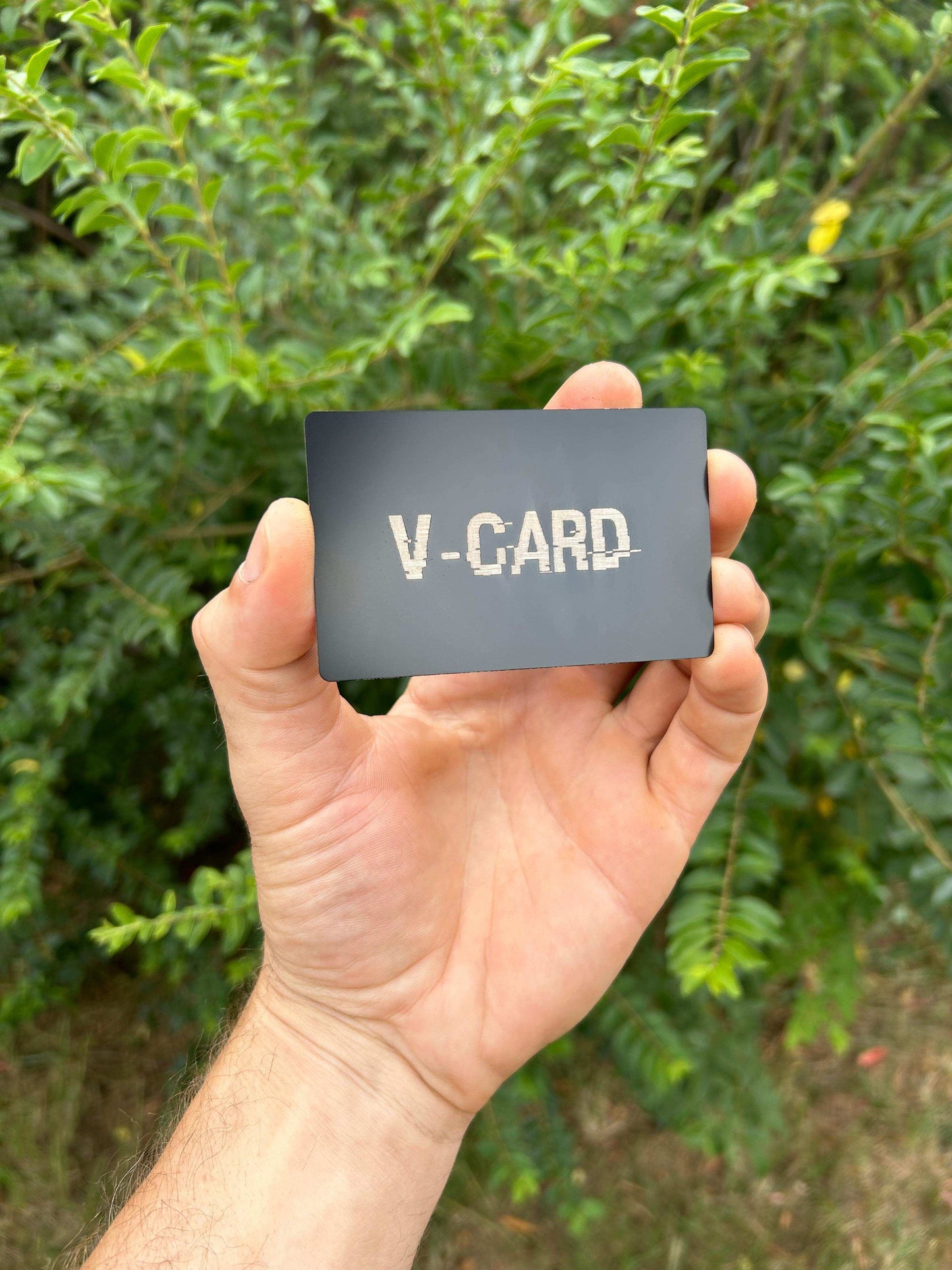 Hand holding a black card with 'V-CARD' text against a green leafy background