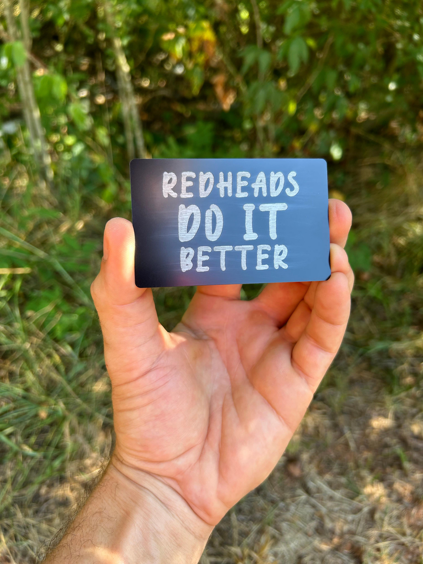 Hand holding a blue card with 'Redheads Do It Better' text against a natural background
