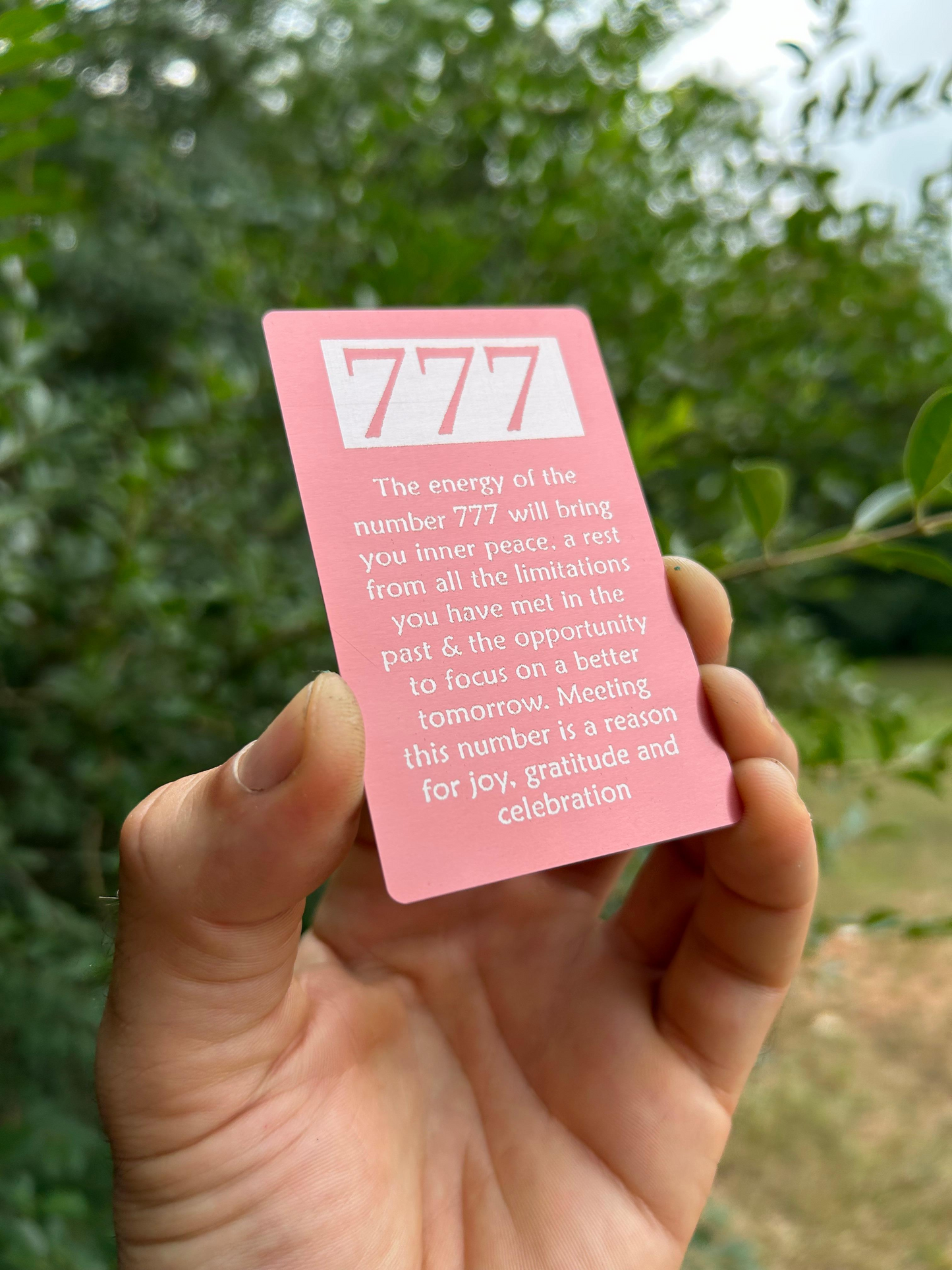 Hand holding a laser engraved numerology card with '777' and text against a green leafy background