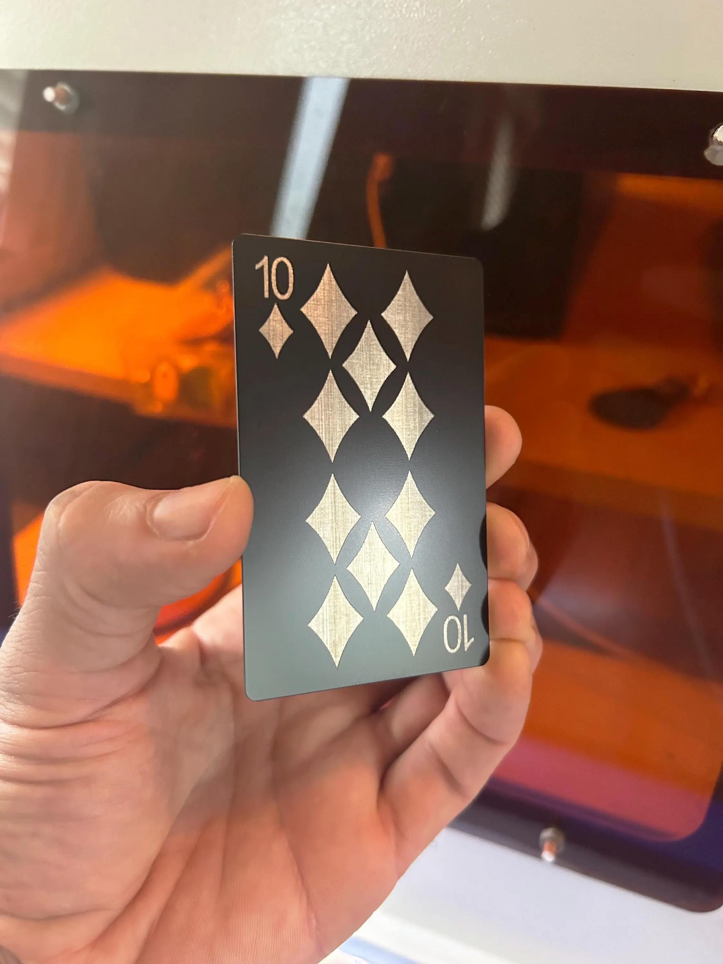 Hand holding a black and silver laser engraved ten of diamonds playing card in front of a blurred machine