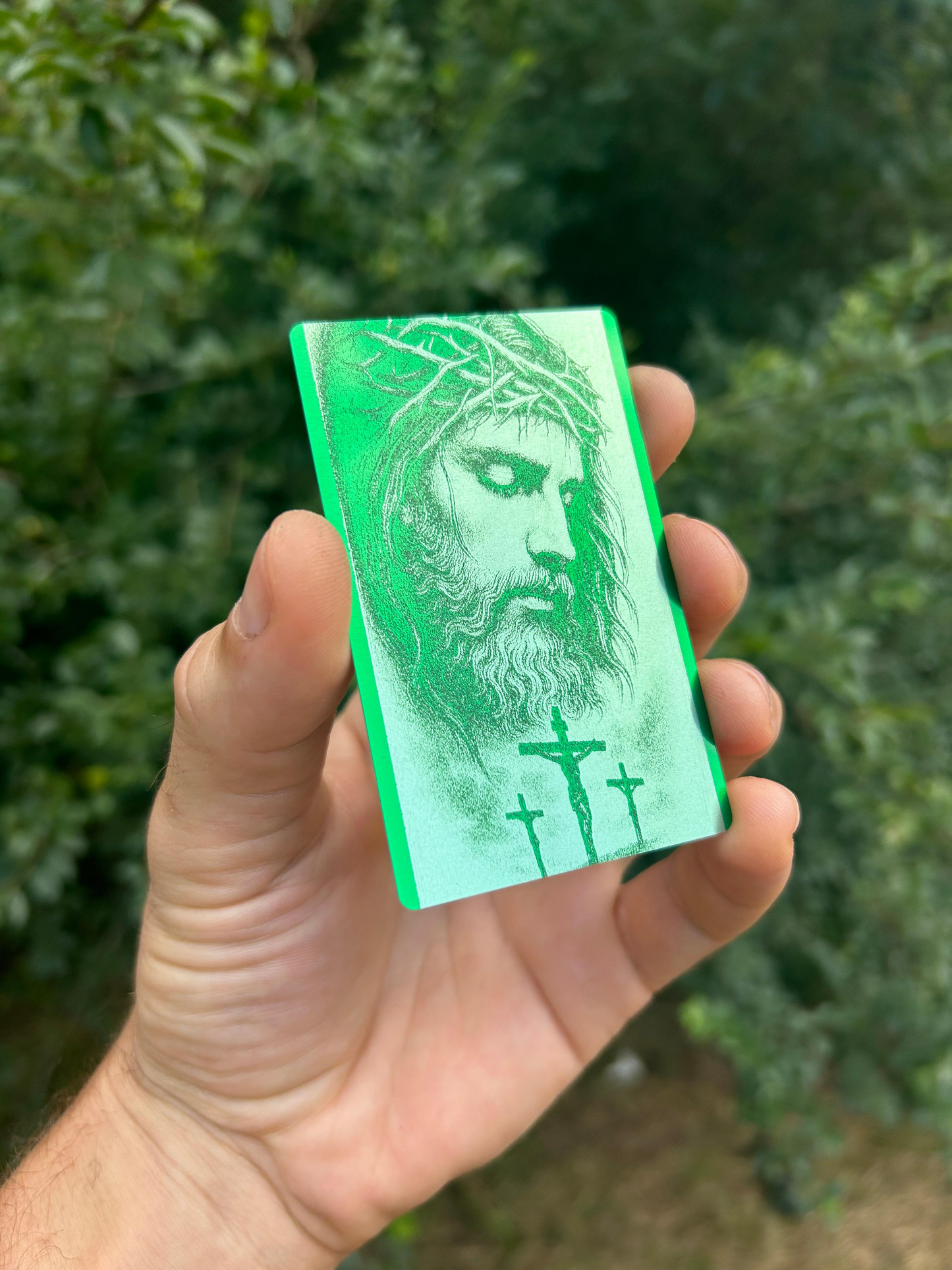 Hand holding a metal green card with a silver engraved crucifix illustration design against a blurred green background