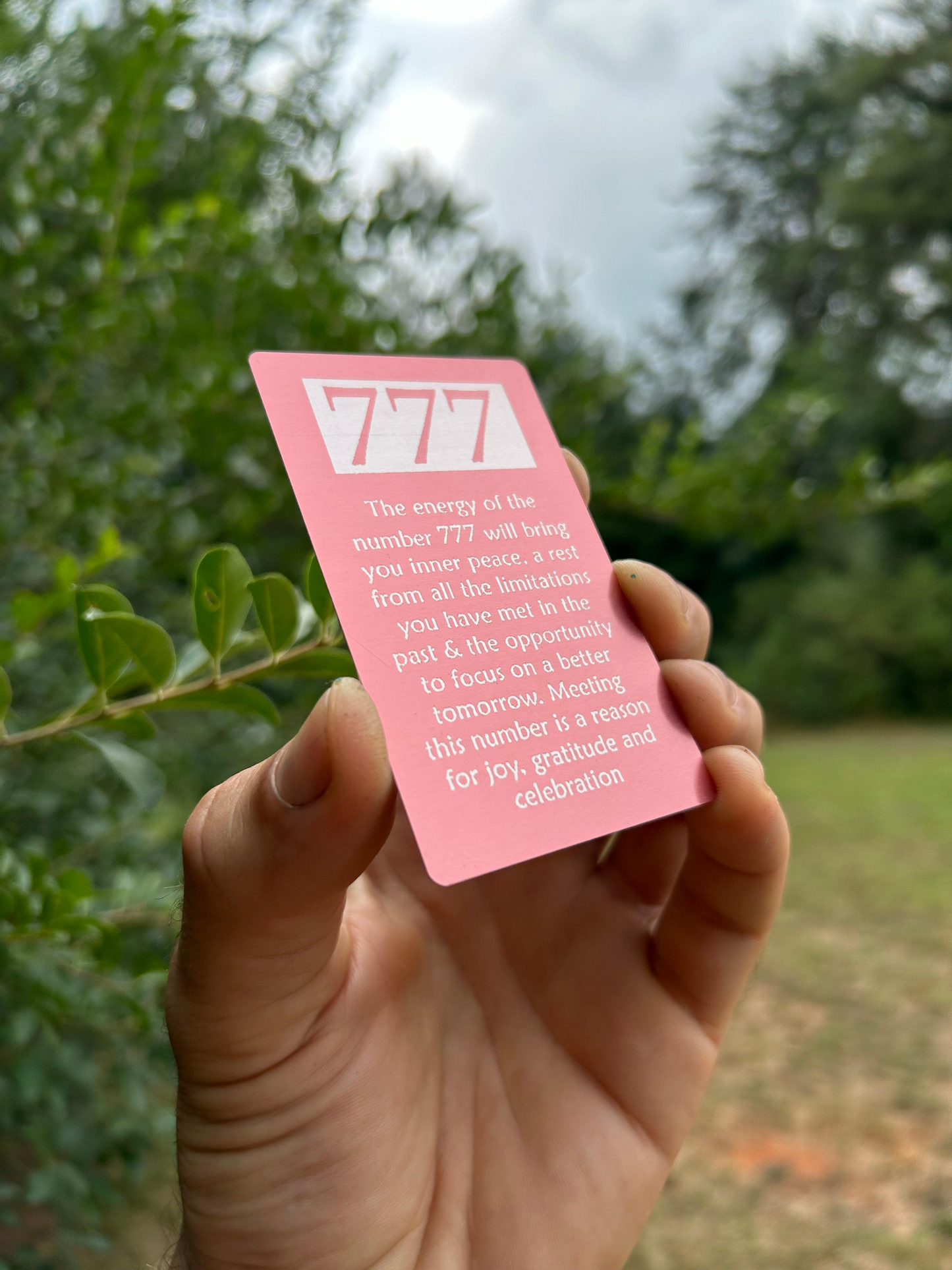 Hand holding a pink card with '777' and text against a blurred green outdoor background
