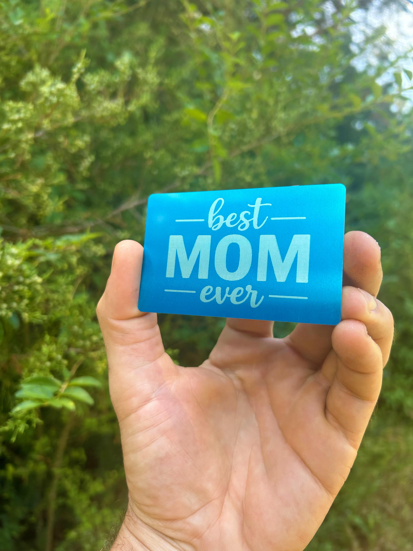 Hand holding a blue/silver metal engraved card with 'best mom ever' text against a green leafy background
