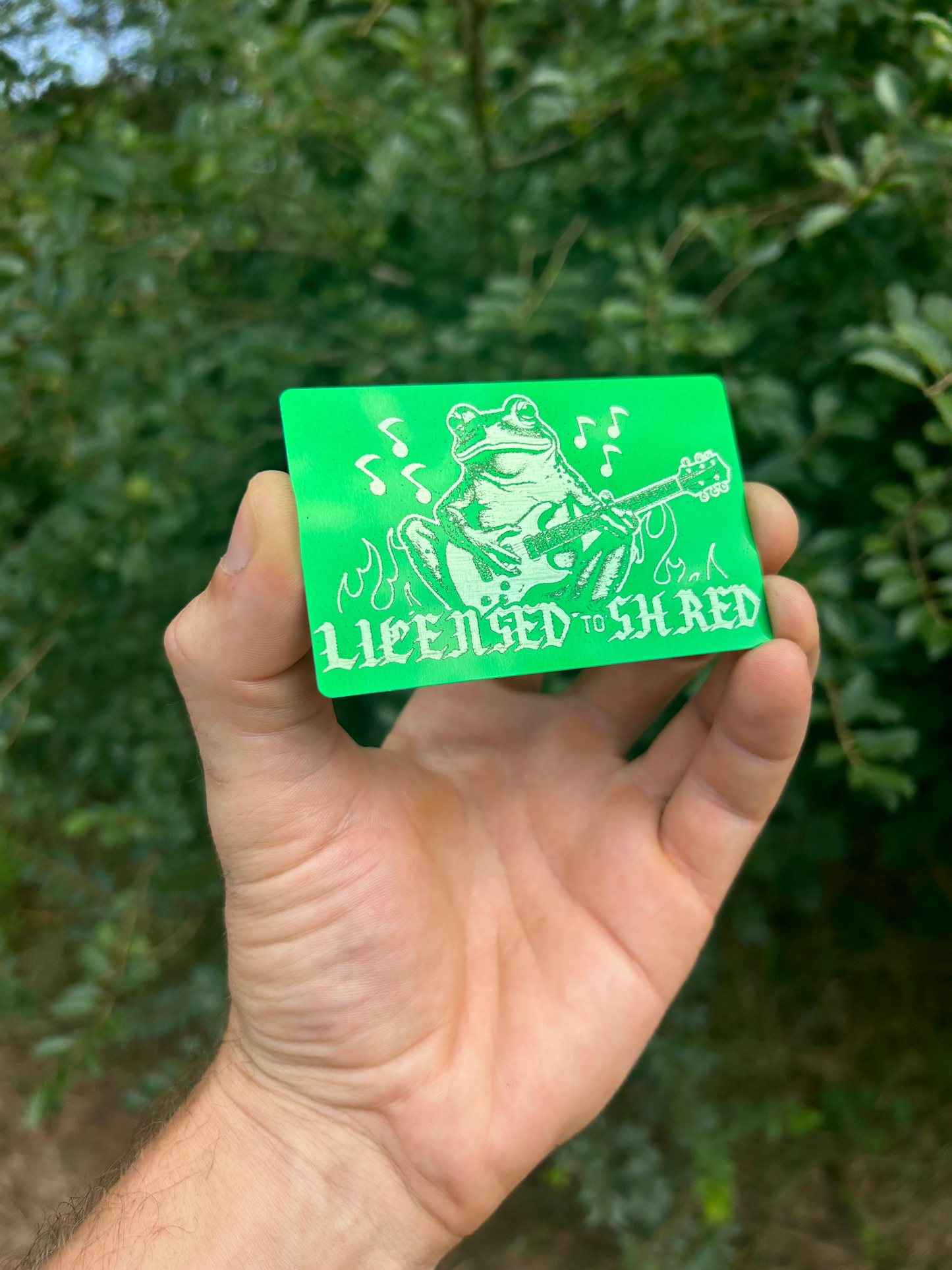 Hand holding a green/silver metal card with a graphic and text against a blurred natural background