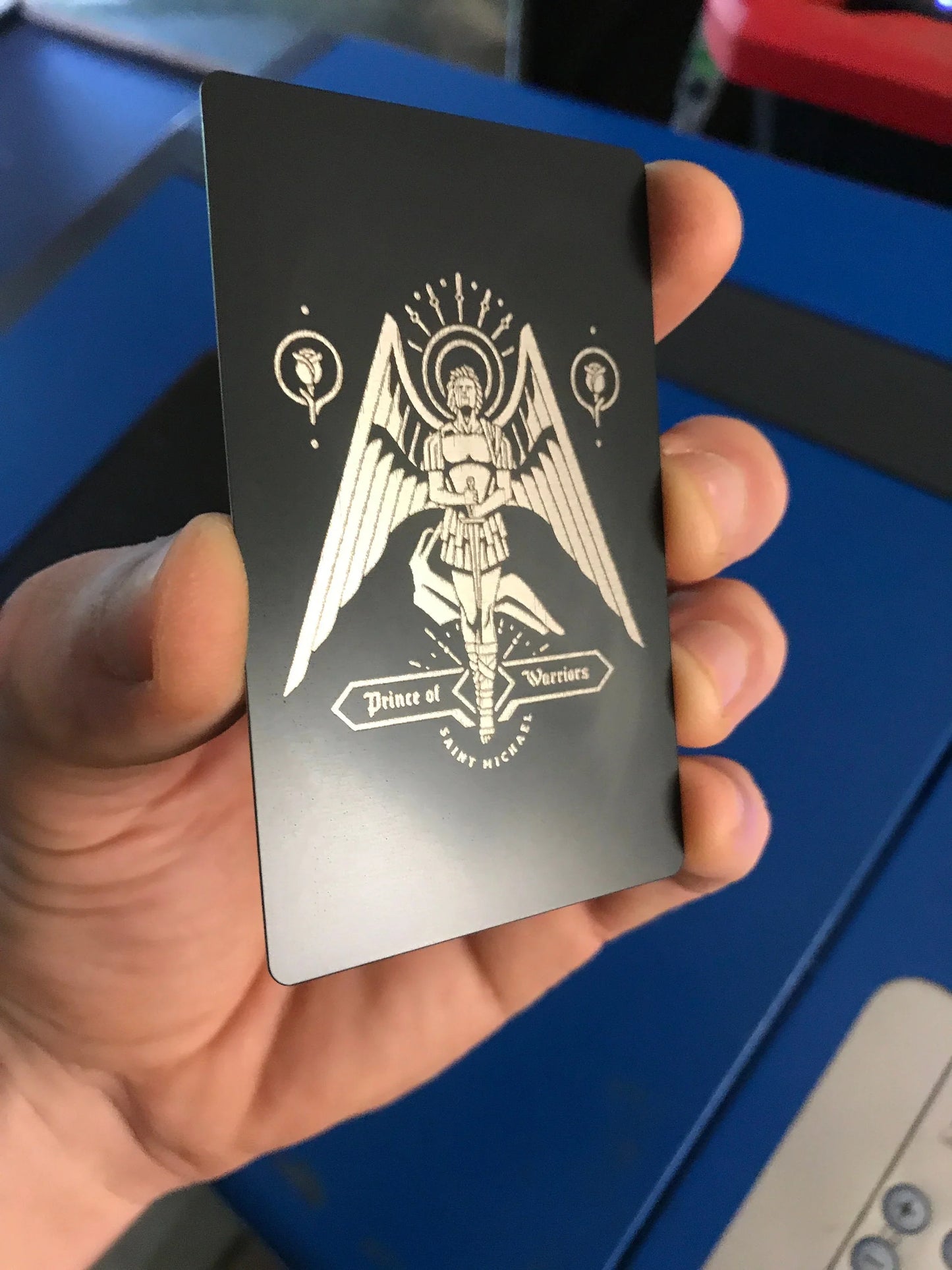 Hand holding a black card with a white St Michael Archangel design against a blue machine background