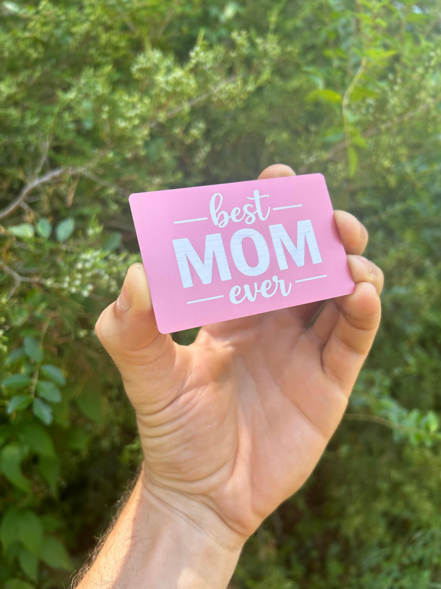 Hand holding a metal rose gold pink card with 'best mom ever' text against a green leafy background