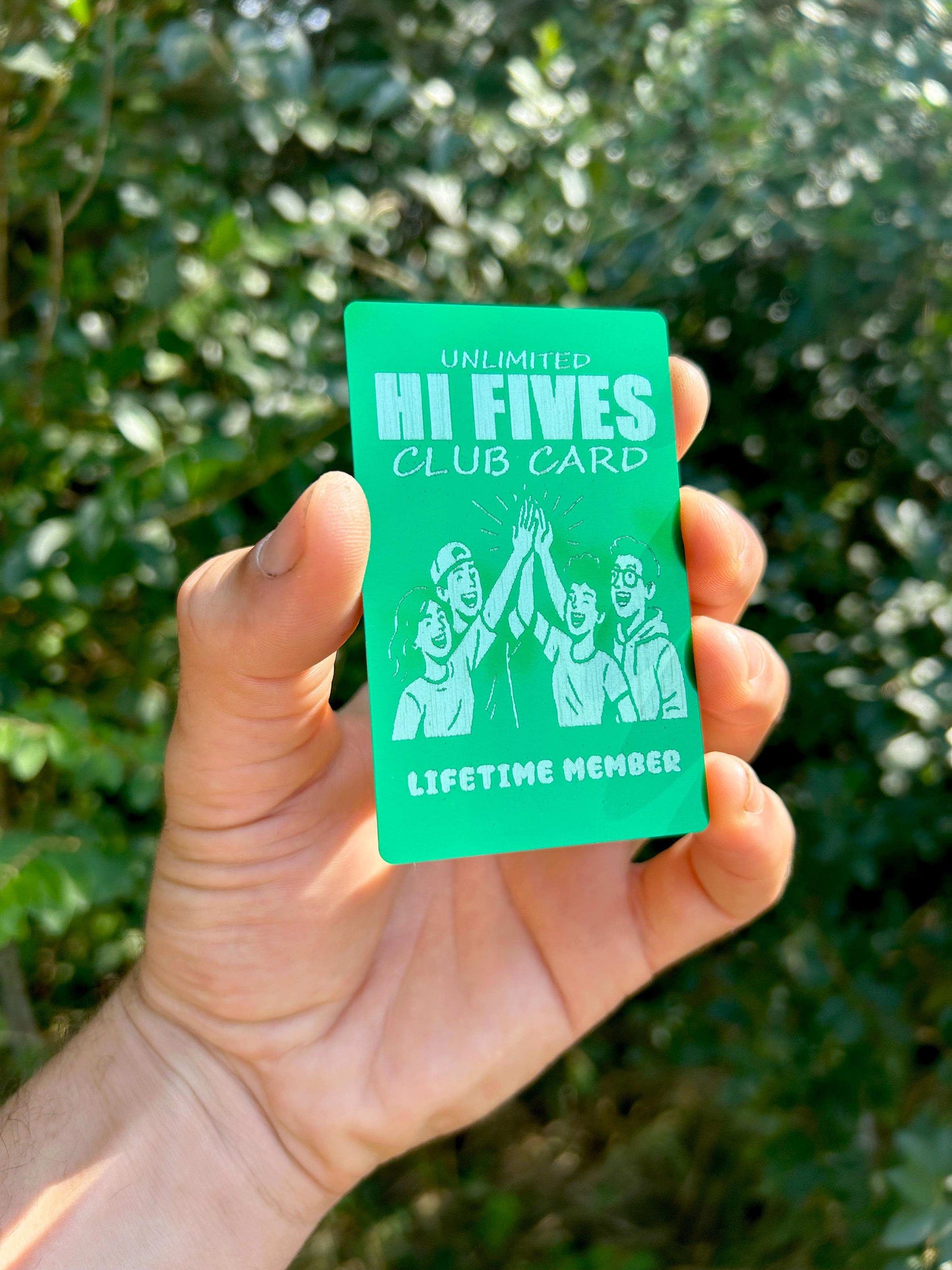 Hand holding a fun, metal, green/silver 'Hi Fives Club Card' with a blurred green leafy background