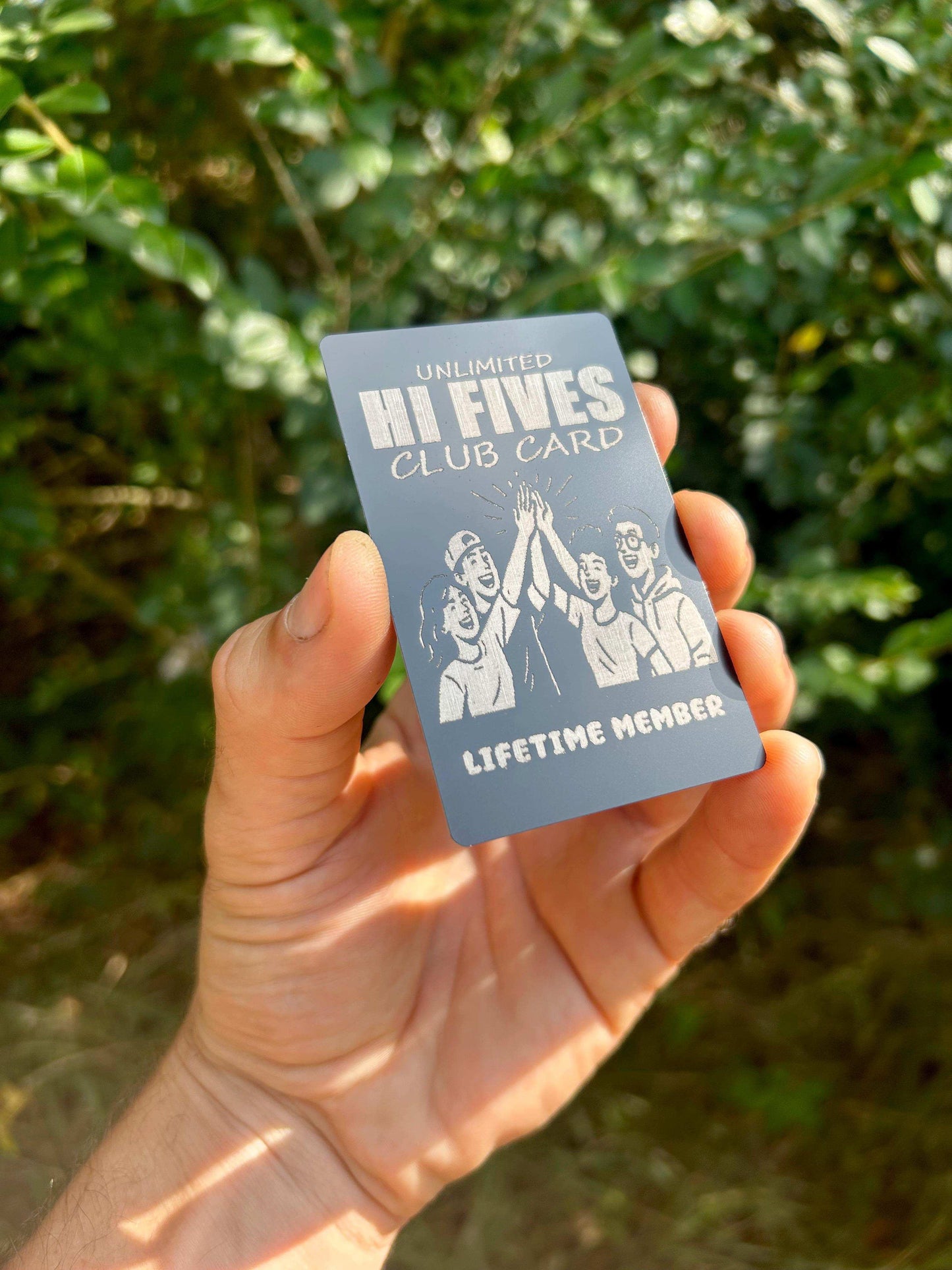 Hand holding a metal 'Hi Fives Club Card' engraving with a green background