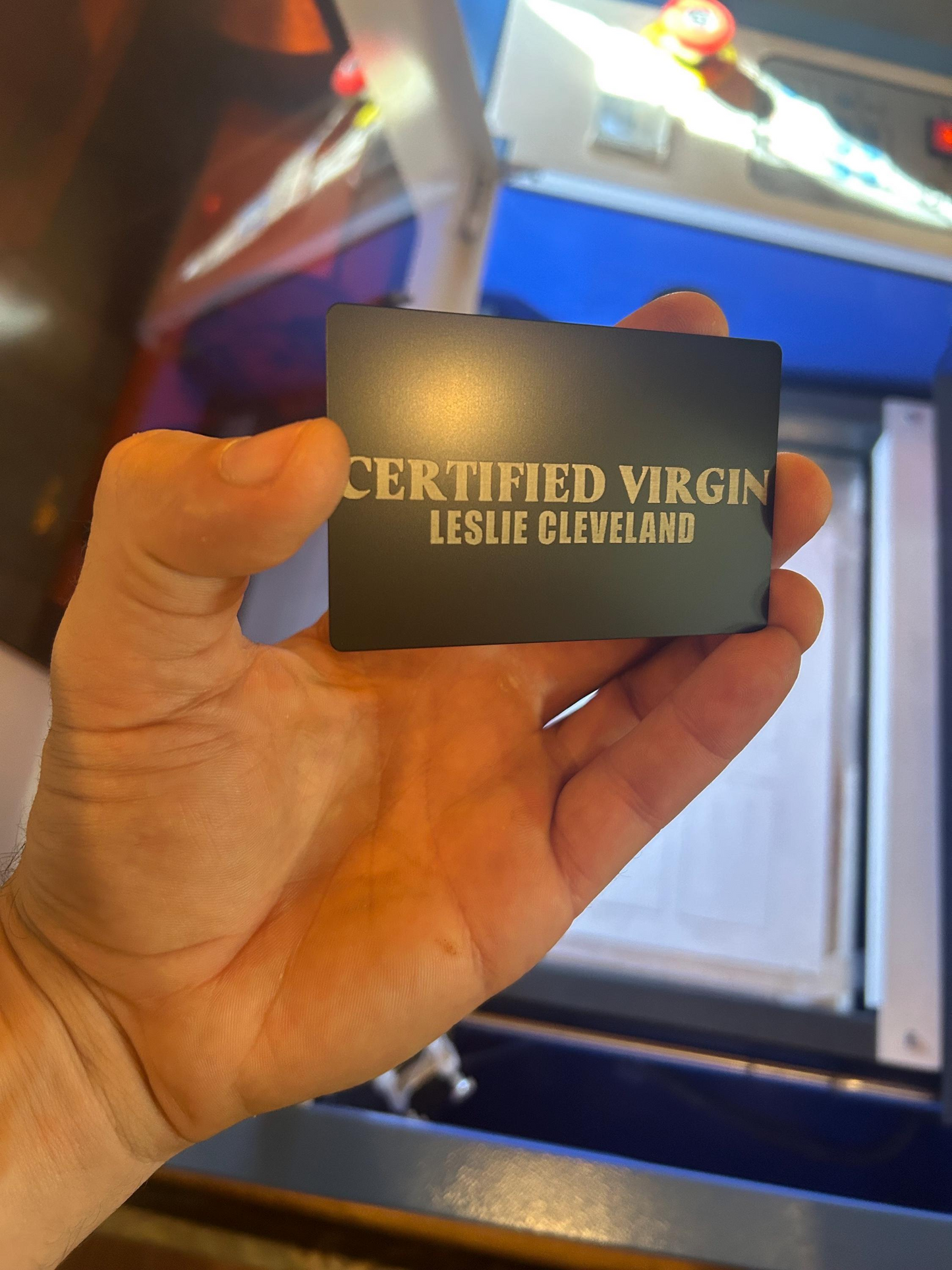 Hand holding a card with 'Certified Virgin' text with a custom name laser engraved against a blurred background