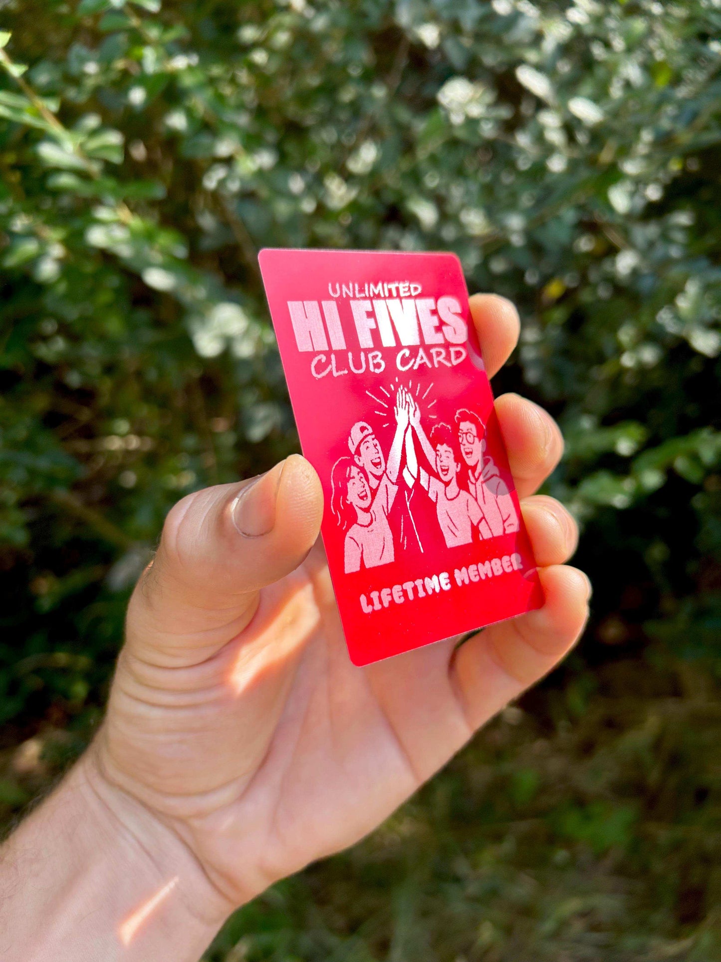 Hand holding a funny metal red 'Unlimited Hi Fives Club Card' with a leafy background