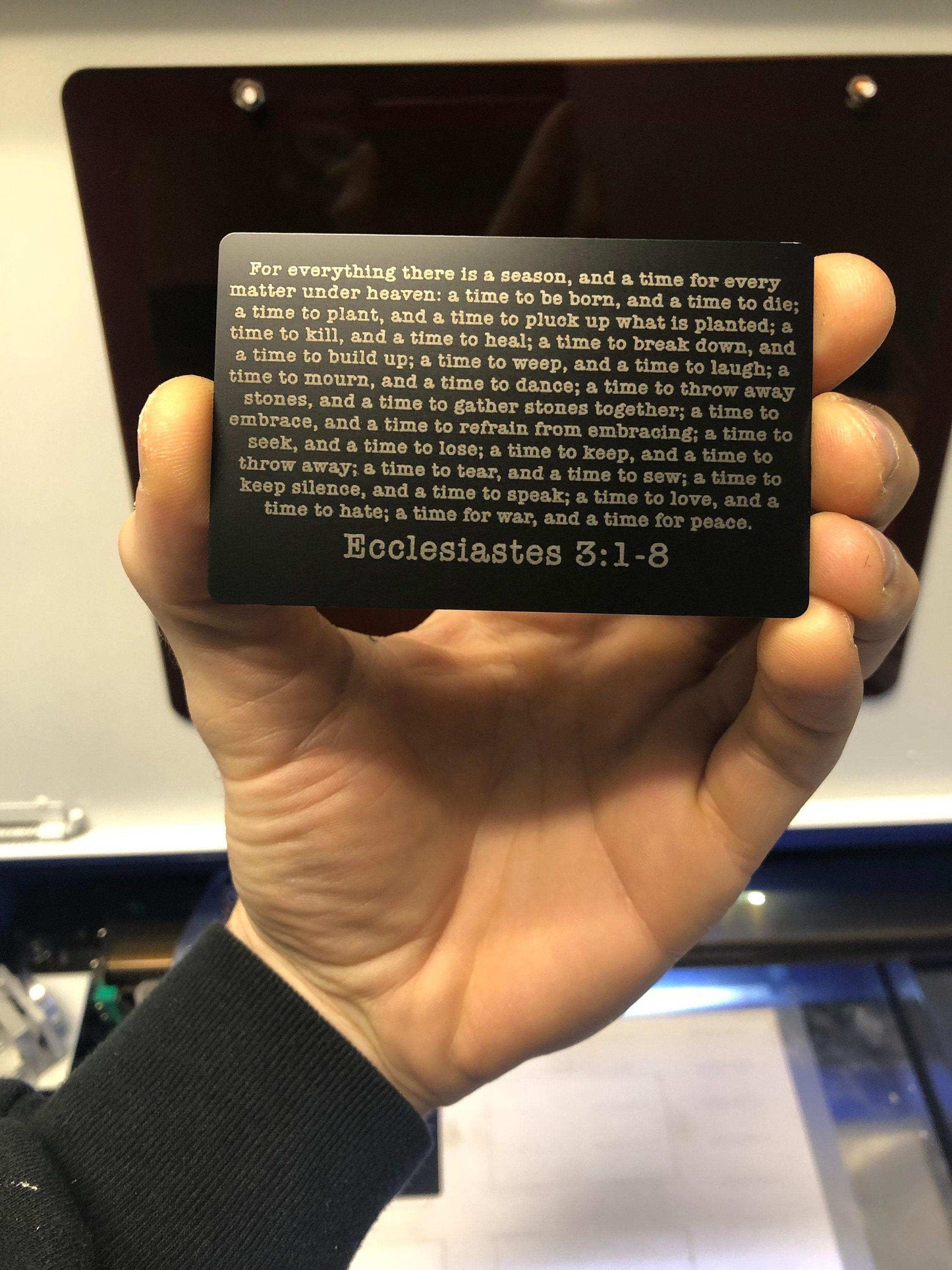 Hand holding a laser engraved metal bible verse card in black/silver with text and 'Ecclesiastes 3:1-8' on it.