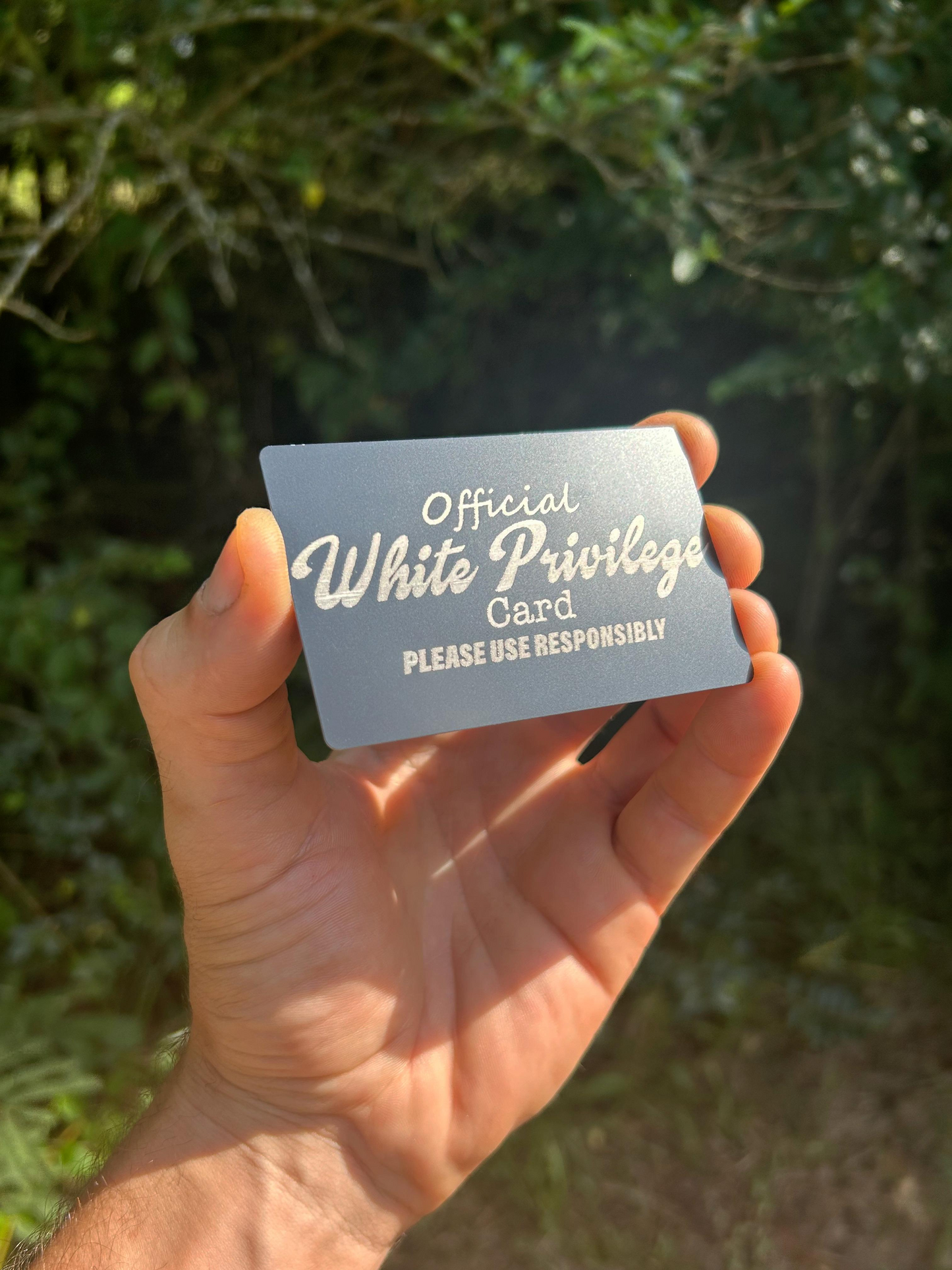 Hand holding a laser engraved gift with 'Official White Privilege Card' text against a blurred natural background