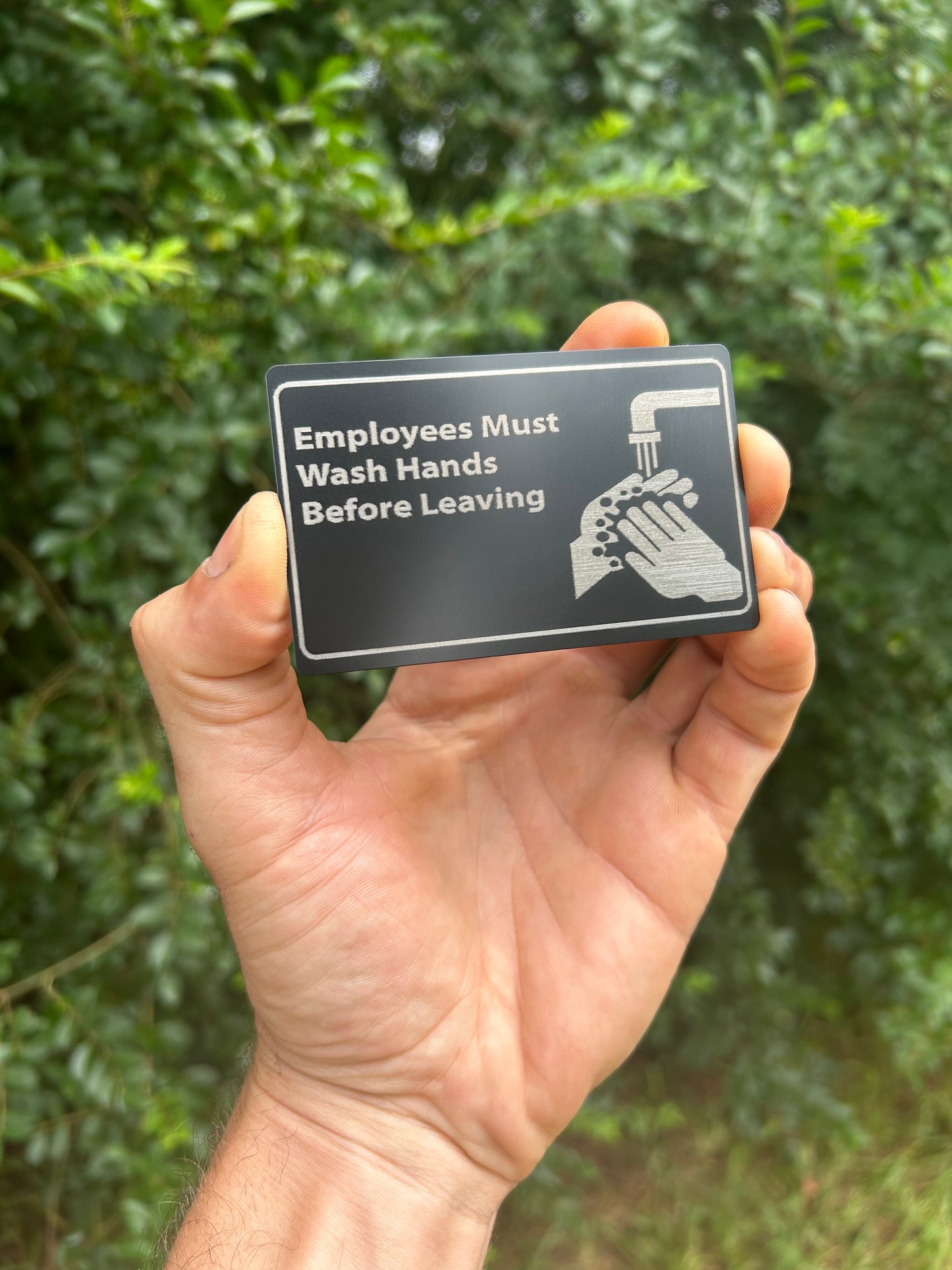Hand holding a sign with text 'Employees Must Wash Hands Before Leaving' against a green leafy background.