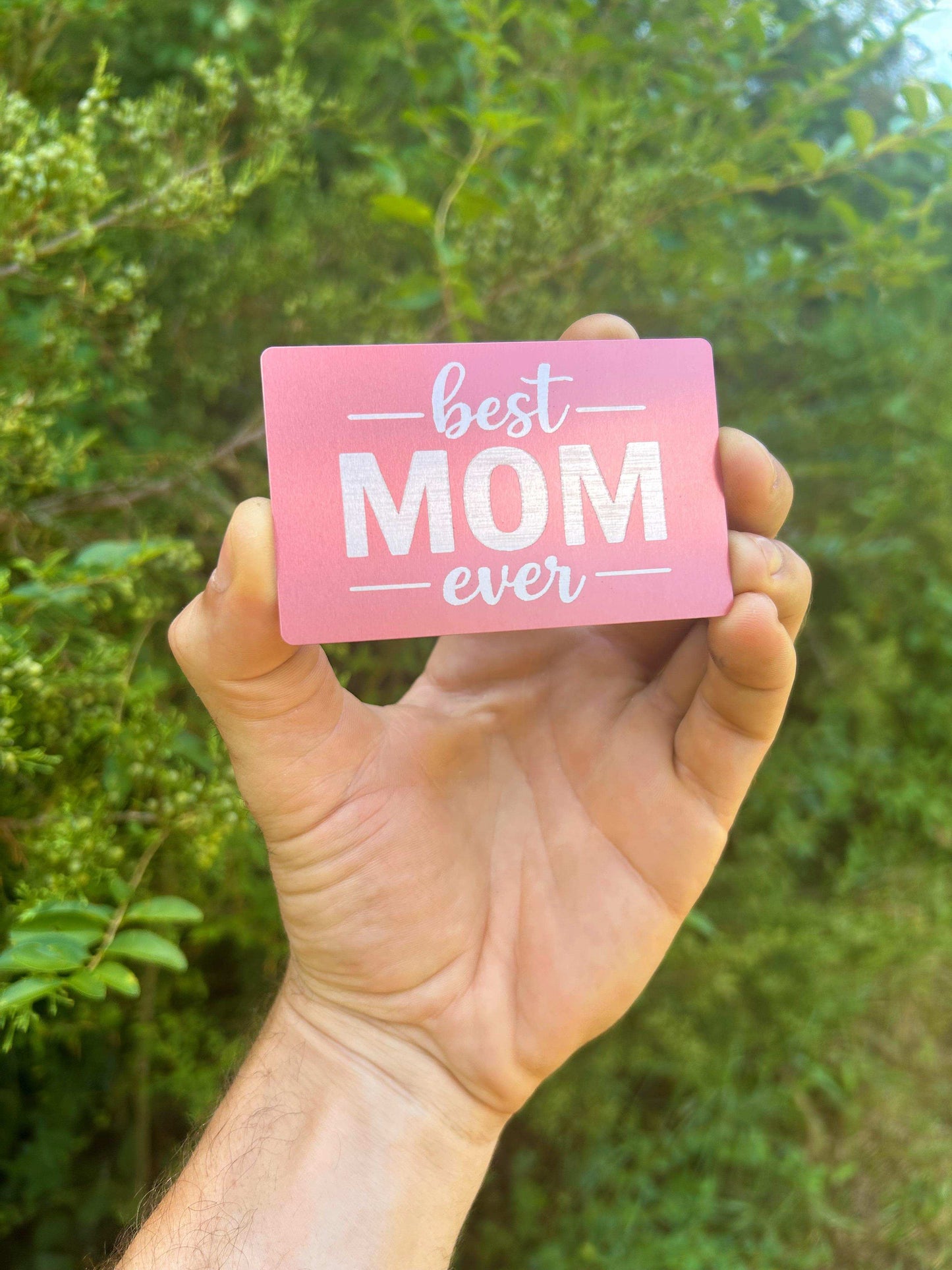 Hand holding a metal greeting card with 'best mom ever' text against a green leafy background