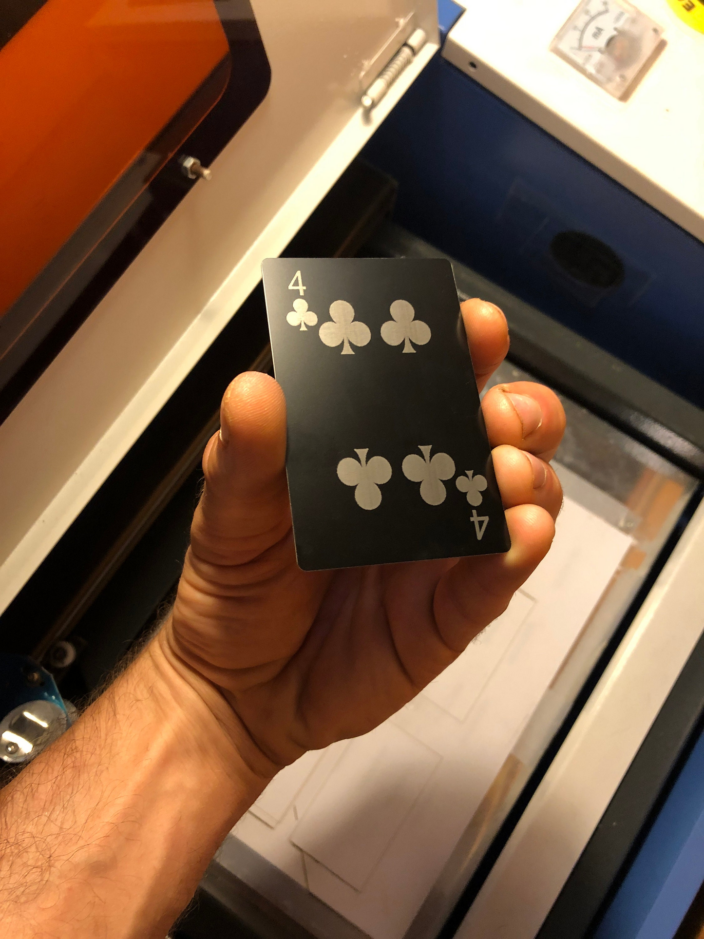 Hand holding a black metal playing card with silver laser engraved Four of Clubs symbols in front of a machine.