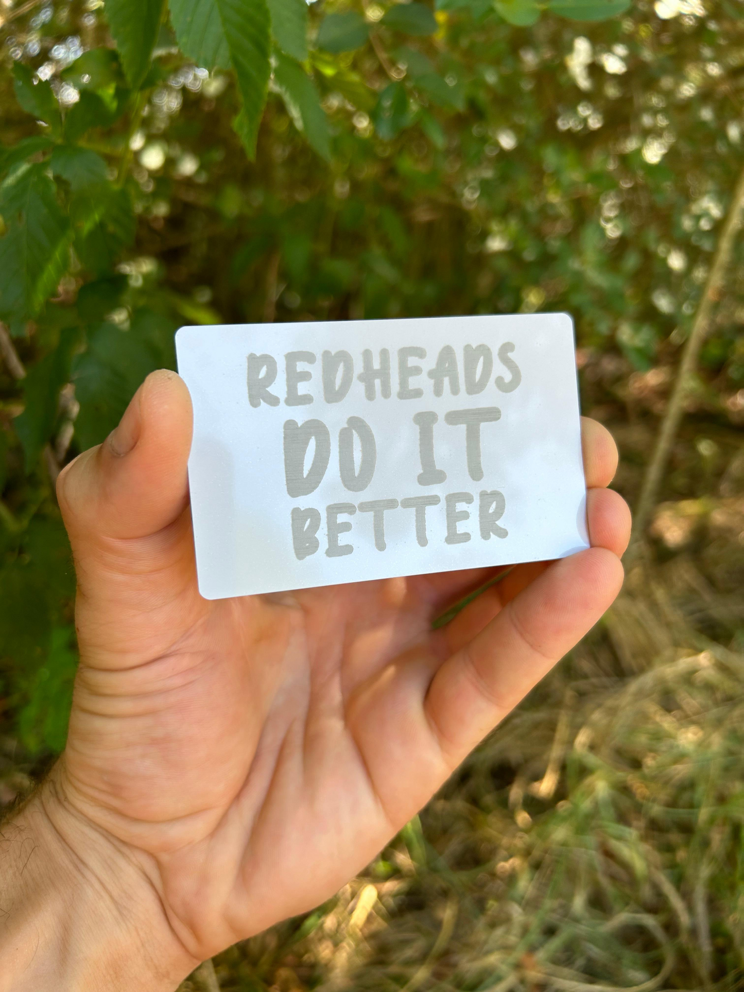 Hand holding a card with 'Redheads Do It Better' text against a natural background