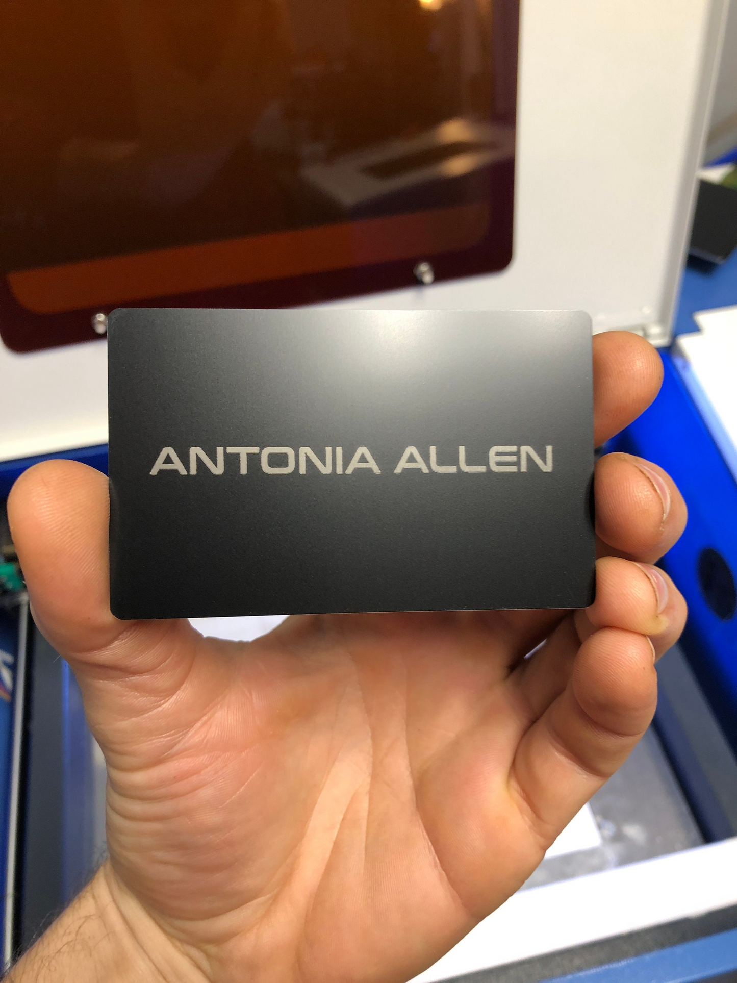 Hand holding a metal black/silver card with 'personalized name' text example engraved against a blurred background