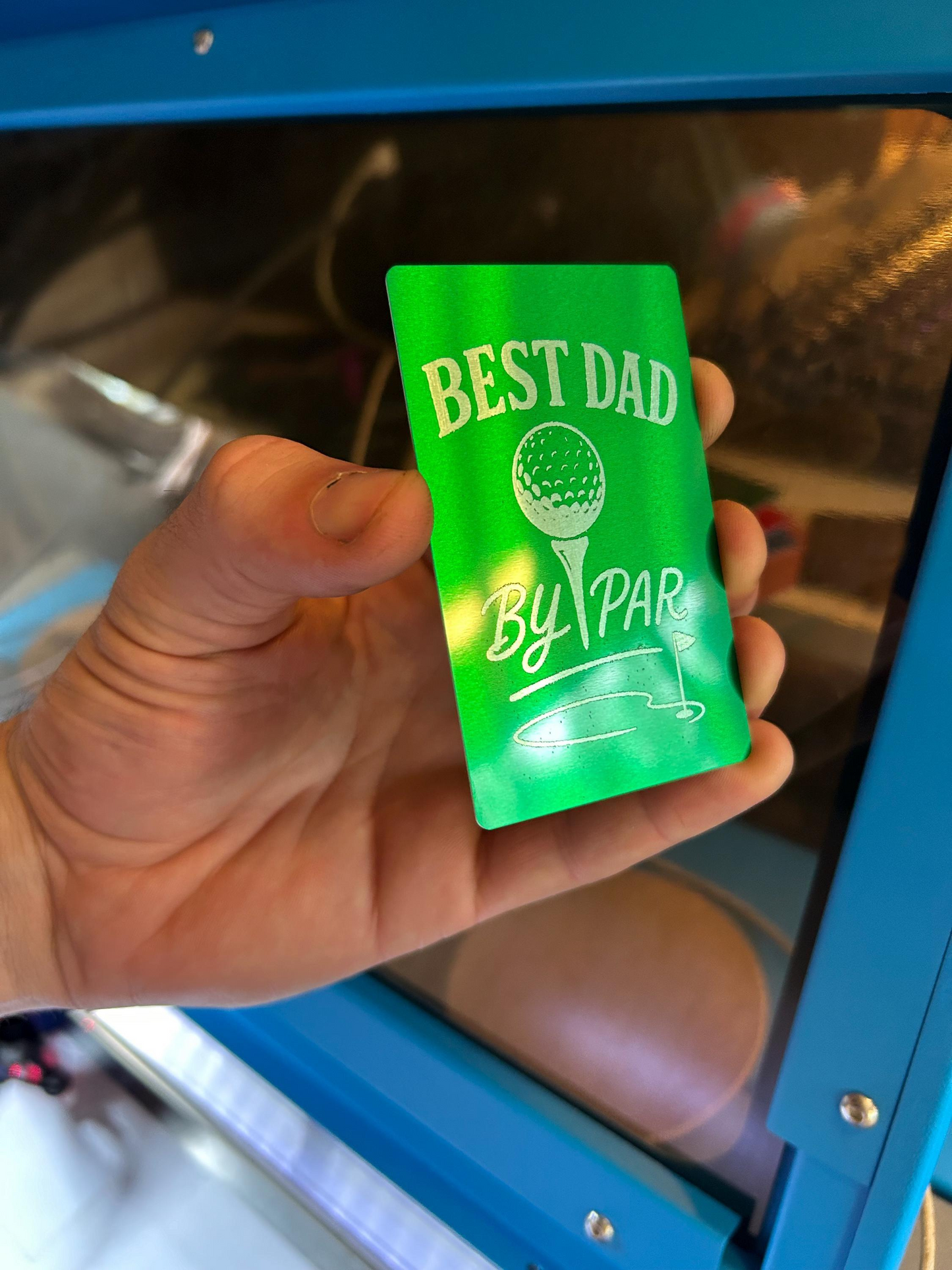 Hand holding a metal silver/green card with 'Best Dad By Par' text against a blurred background