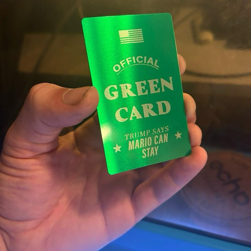 Hand holding a metal "official" green card with custom name text, against a blurred background