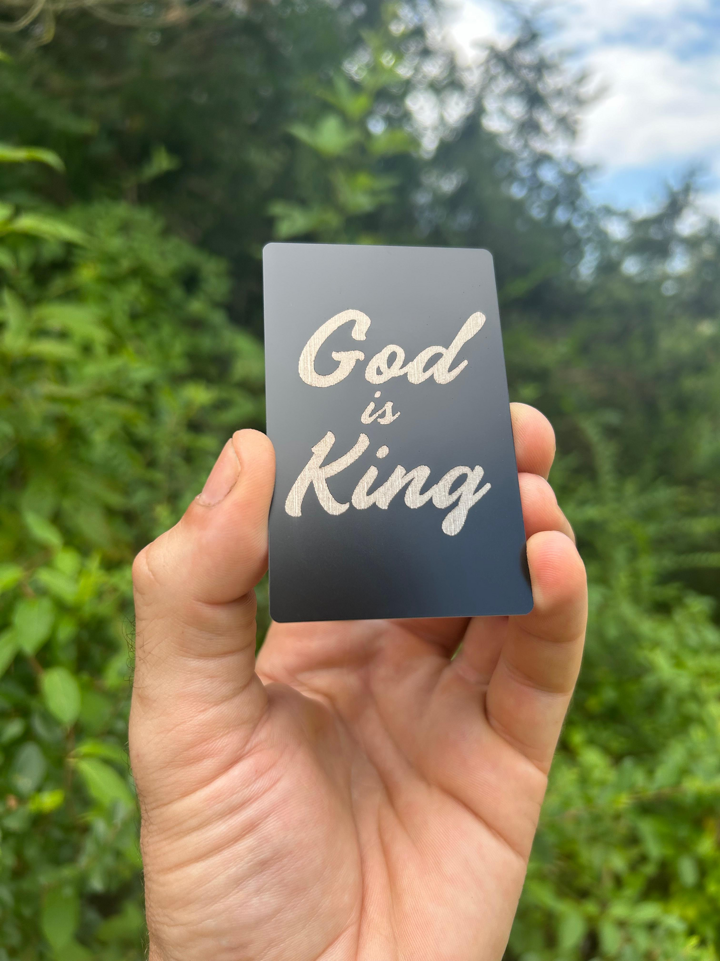 Hand holding a metal laser engraved keepsake card with 'God is King' text against a green outdoor background