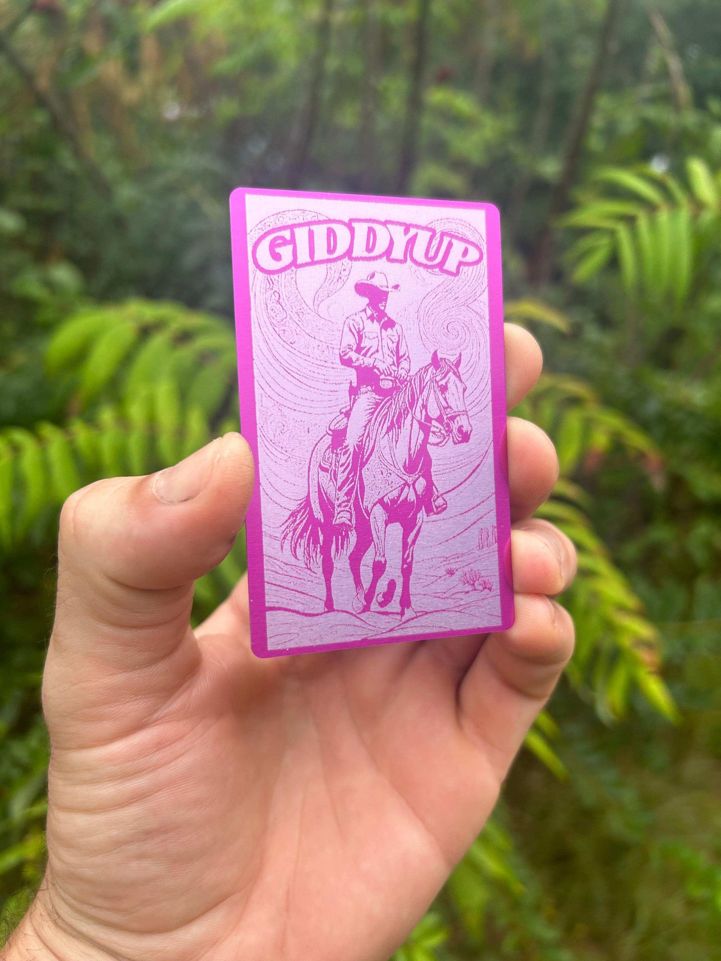Hand holding a purple/silver metal card with a horse and rider illustration and 'Giddy Up' text against a green leafy background.