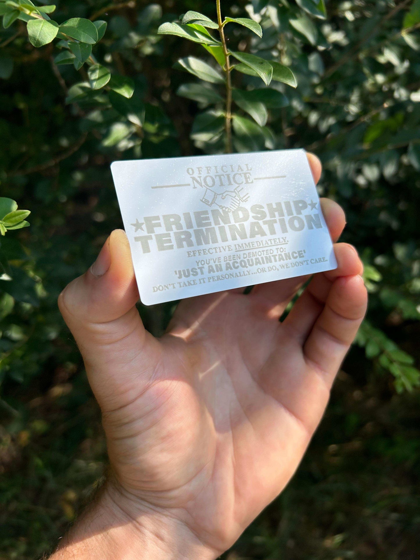Hand holding a metal white/silver "Just An Acquaintance" card with text against a green leafy background