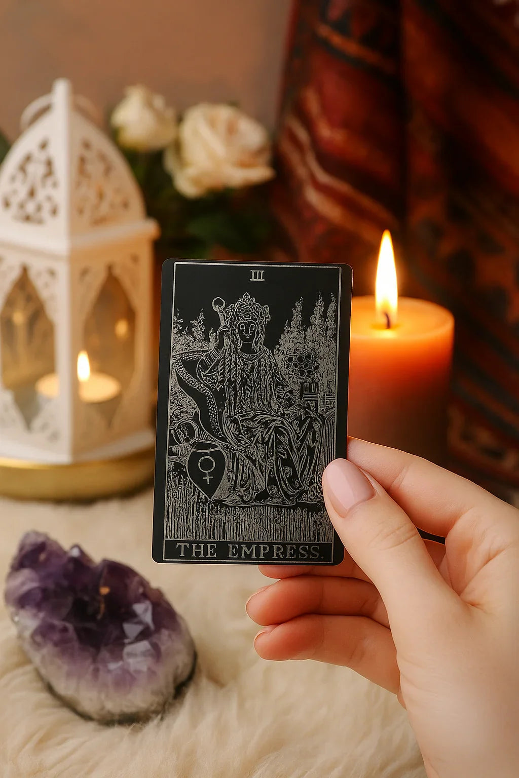 Hand holding an anodized aluminum metal tarot card titled 'The Empress' with candles and crystals in the background