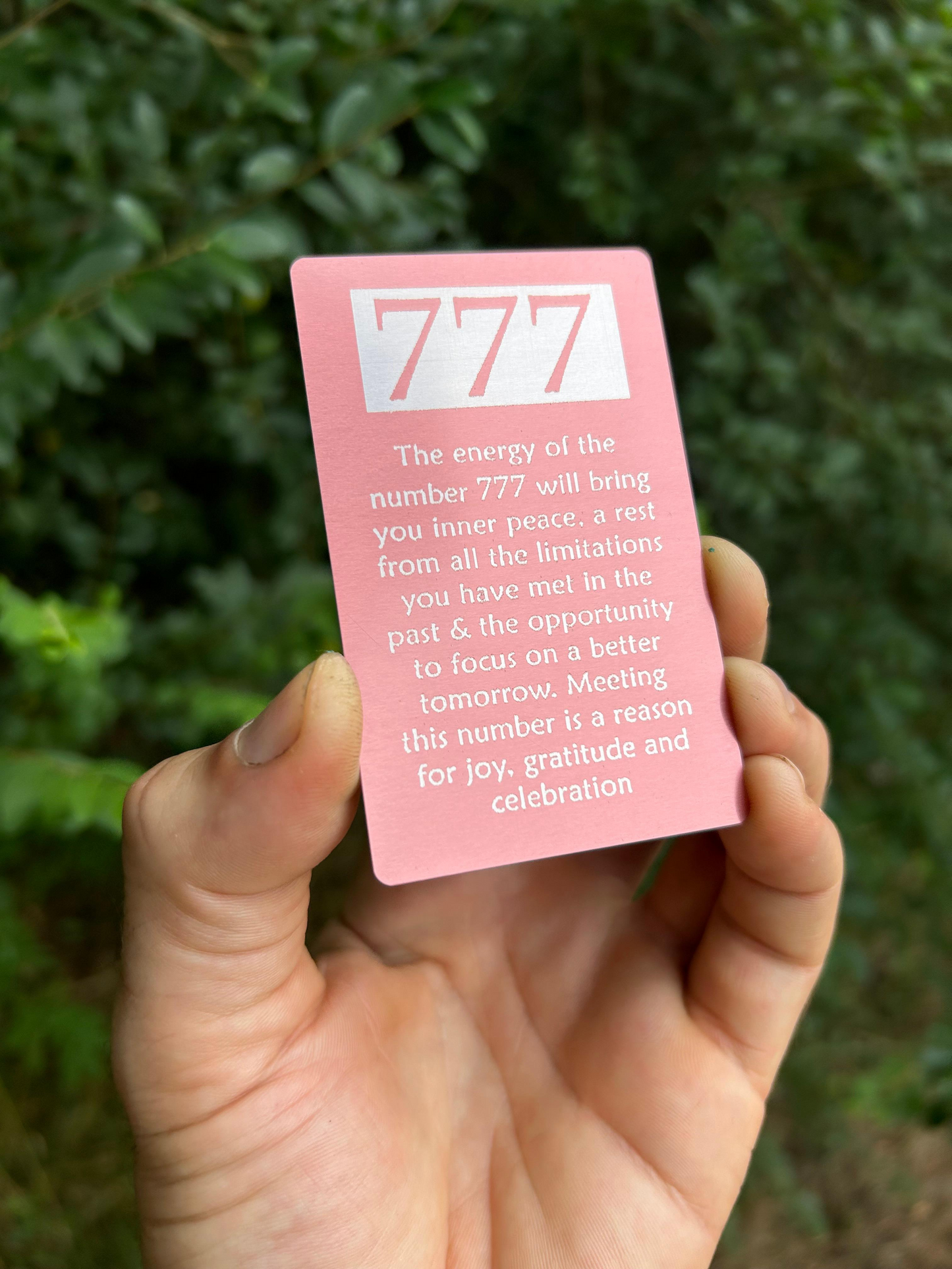 Hand holding a rose gold card with '777' symbol and text against a green leafy background