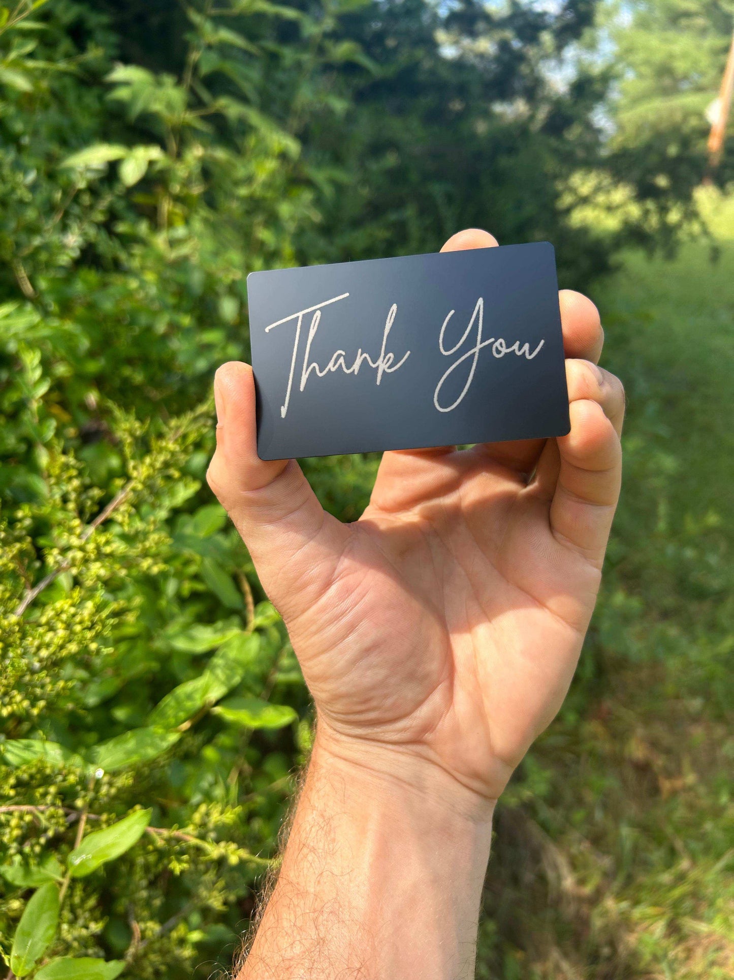 Hand holding a metal black card with 'Thank You' in laser engraved silver cursive text against a green outdoor background