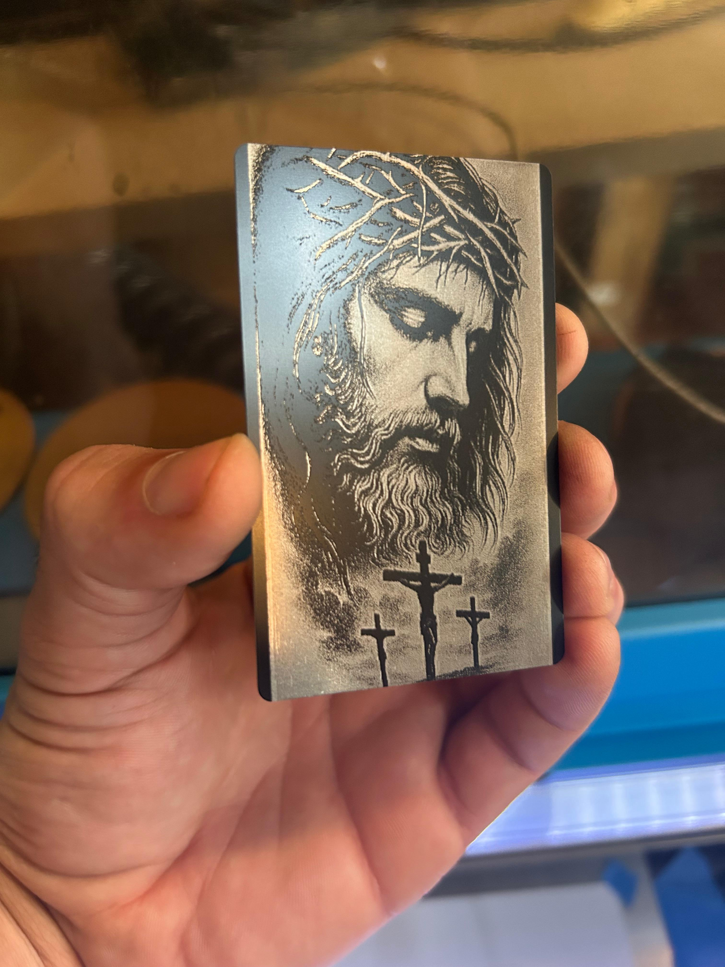 Hand holding a metal bookmark card with engraved image of jesus christ, crown of thorns and the three crosses against an industrial background