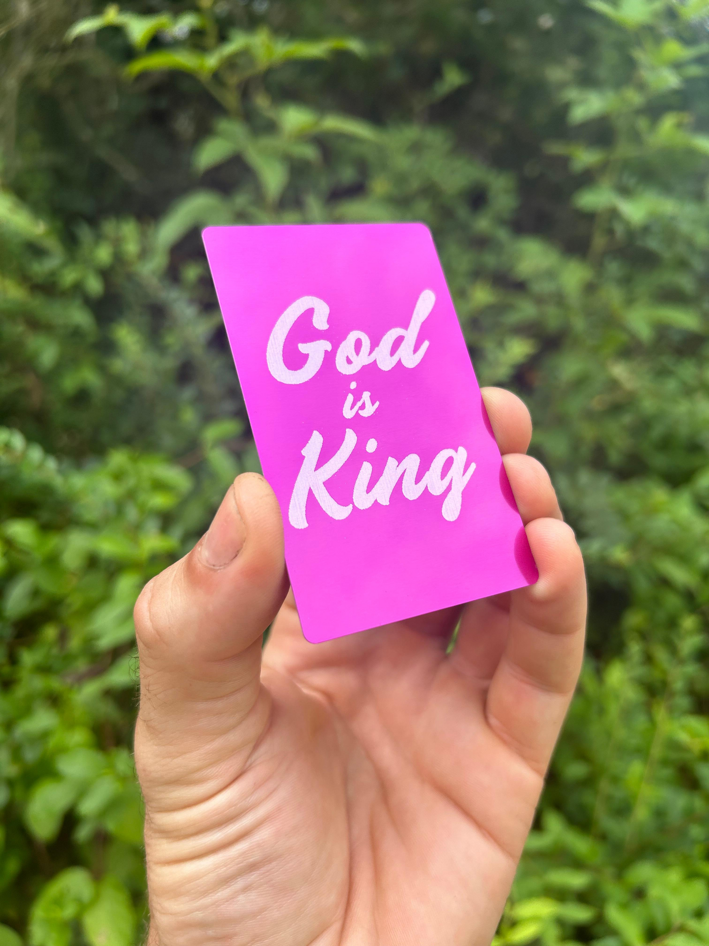 Hand holding a metal purple/silver card with 'God is King' faith inspired text against a green leafy background