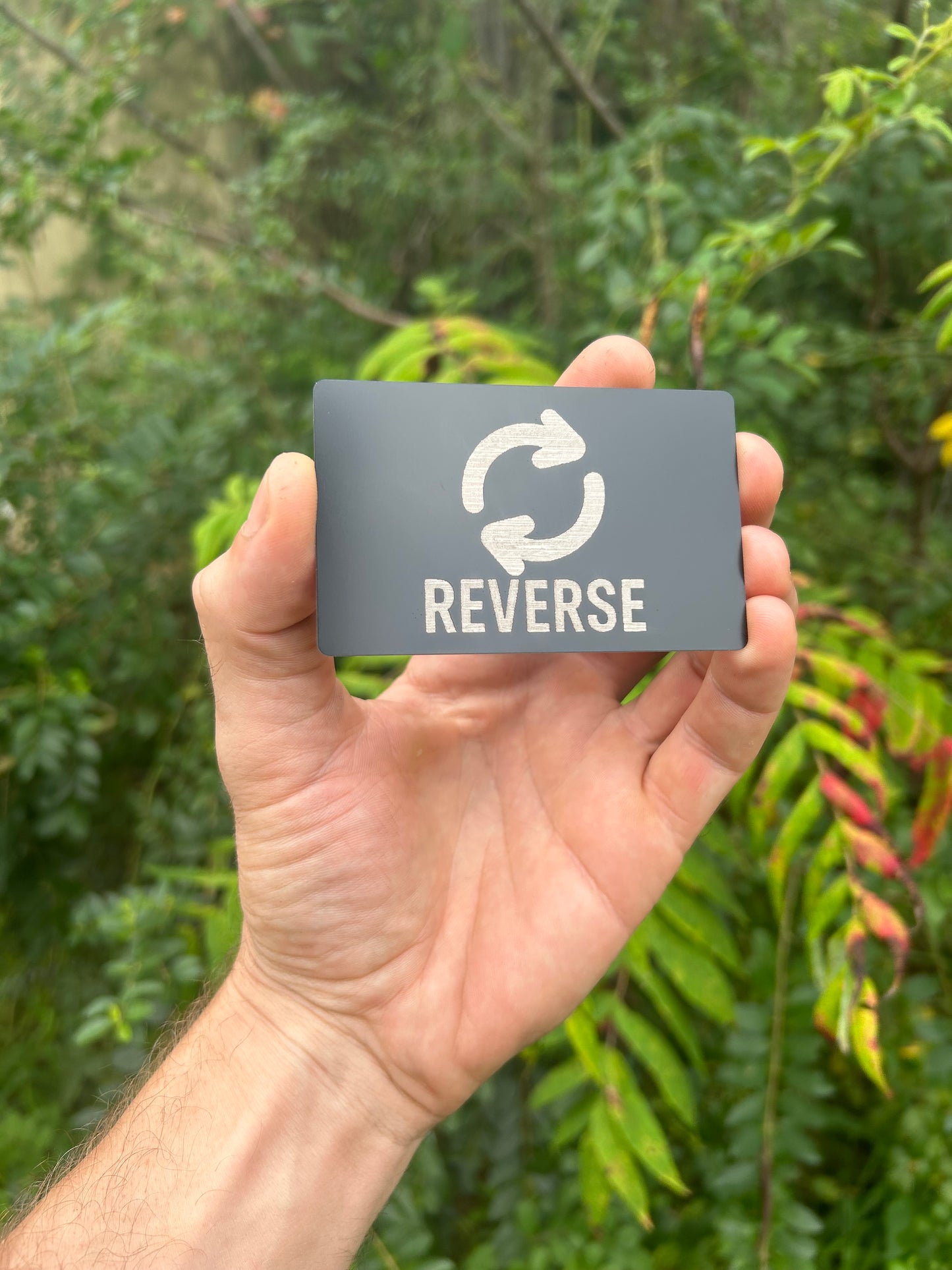 Hand holding a small rectangular metal 'REVERSE' keepsake card and a recycling symbol against a green leafy background