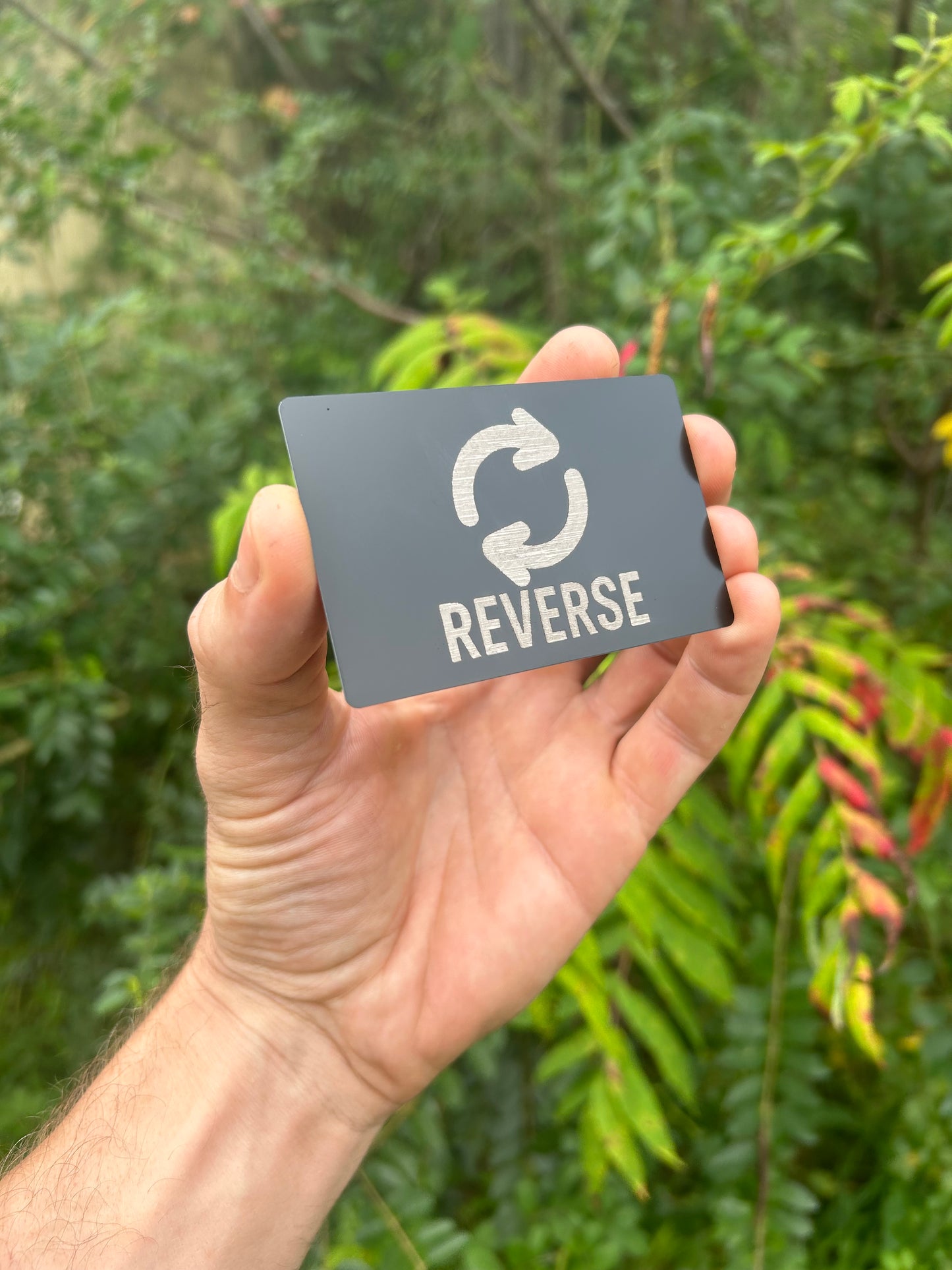 Hand holding a small rectangular card with 'REVERSE' text and emoji symbol against a green leafy background