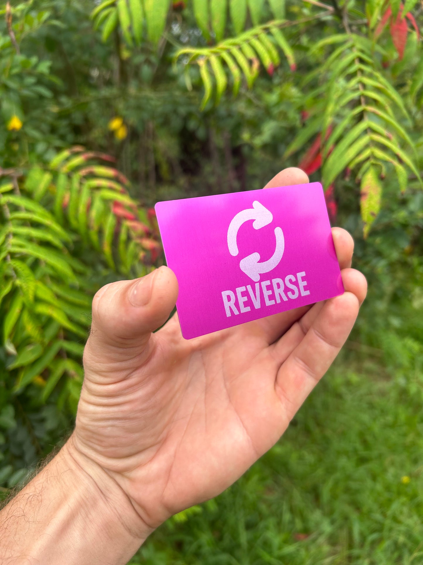 Hand holding a purple/silver metal card with 'REVERSE' text and symbol laser engraved, against a green leafy background