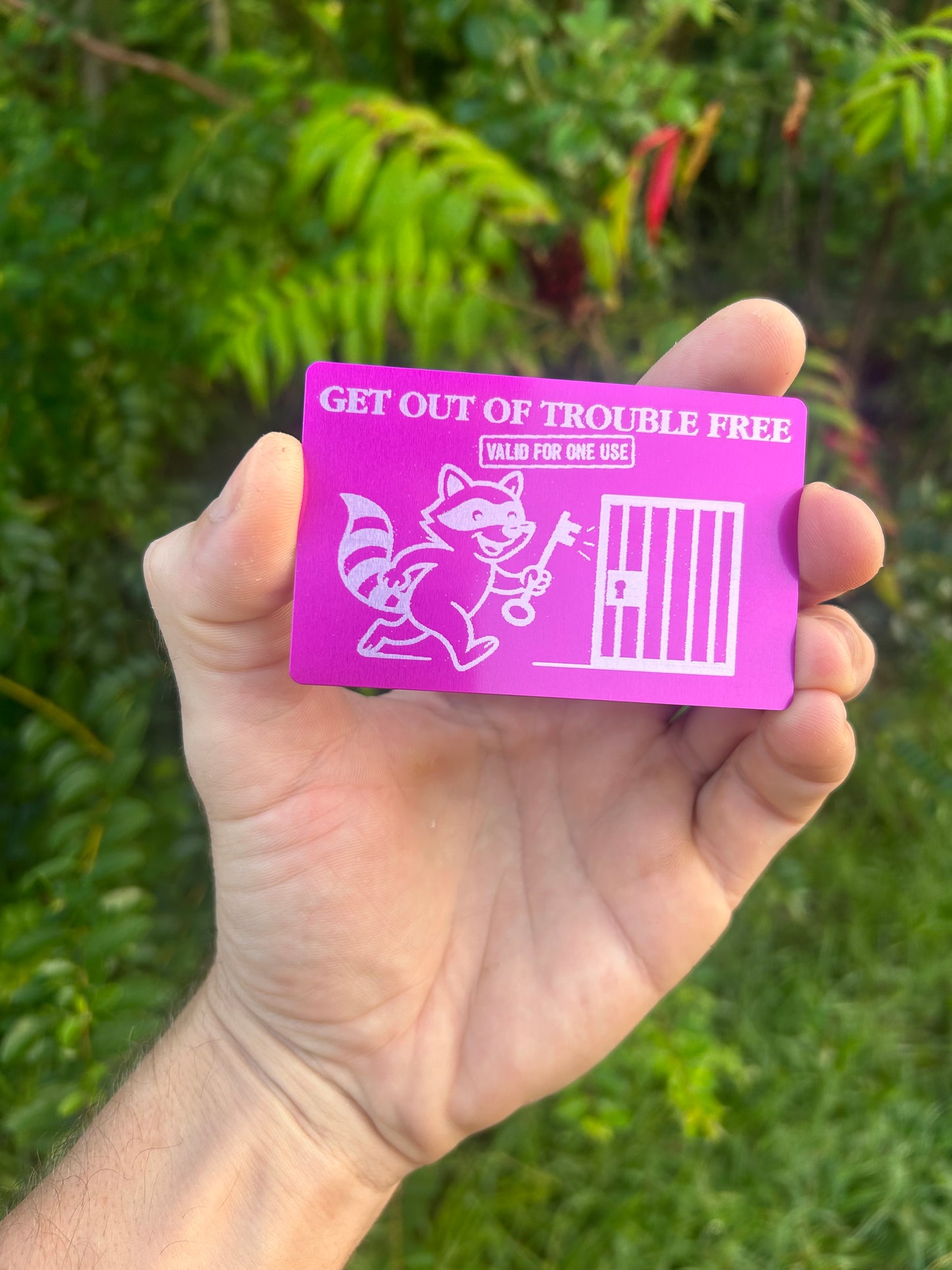 Hand holding a purple metal laser engraved keepsake with "get out of troble free, valid for one use" text and racoon artwork against a green nature background