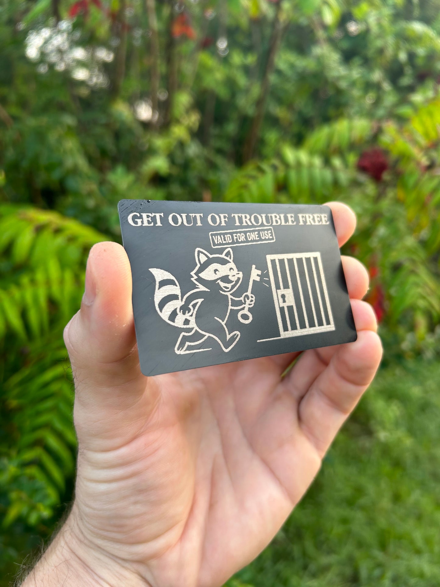 Hand holding a small metal get out of trouble free card with a design and text against a green outdoor background