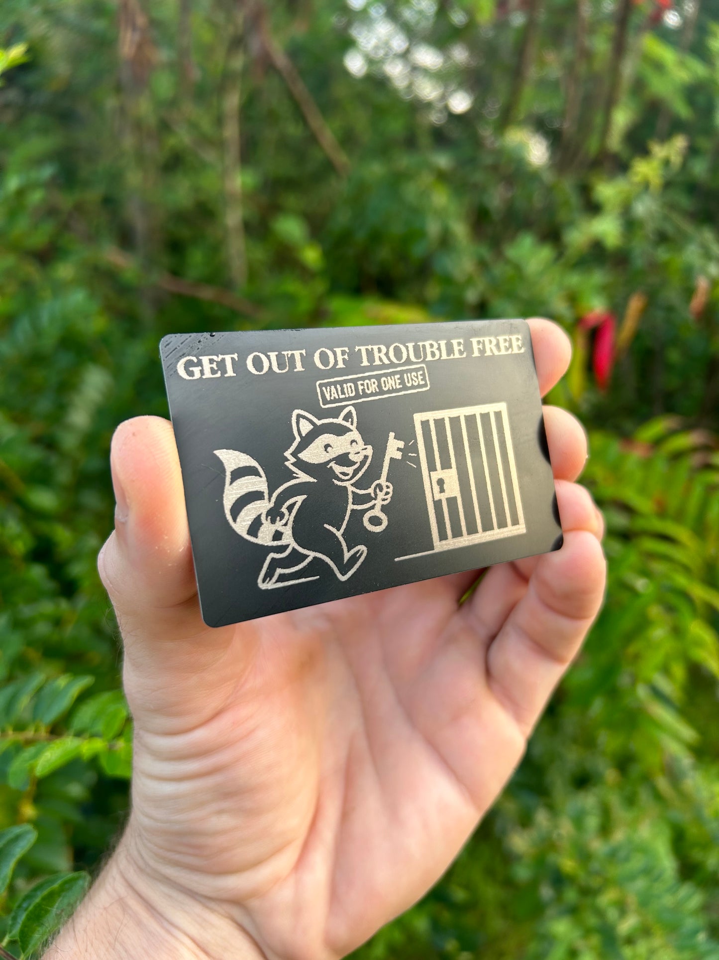 Hand holding a small metal wallet card with text and graphics against a green leafy background