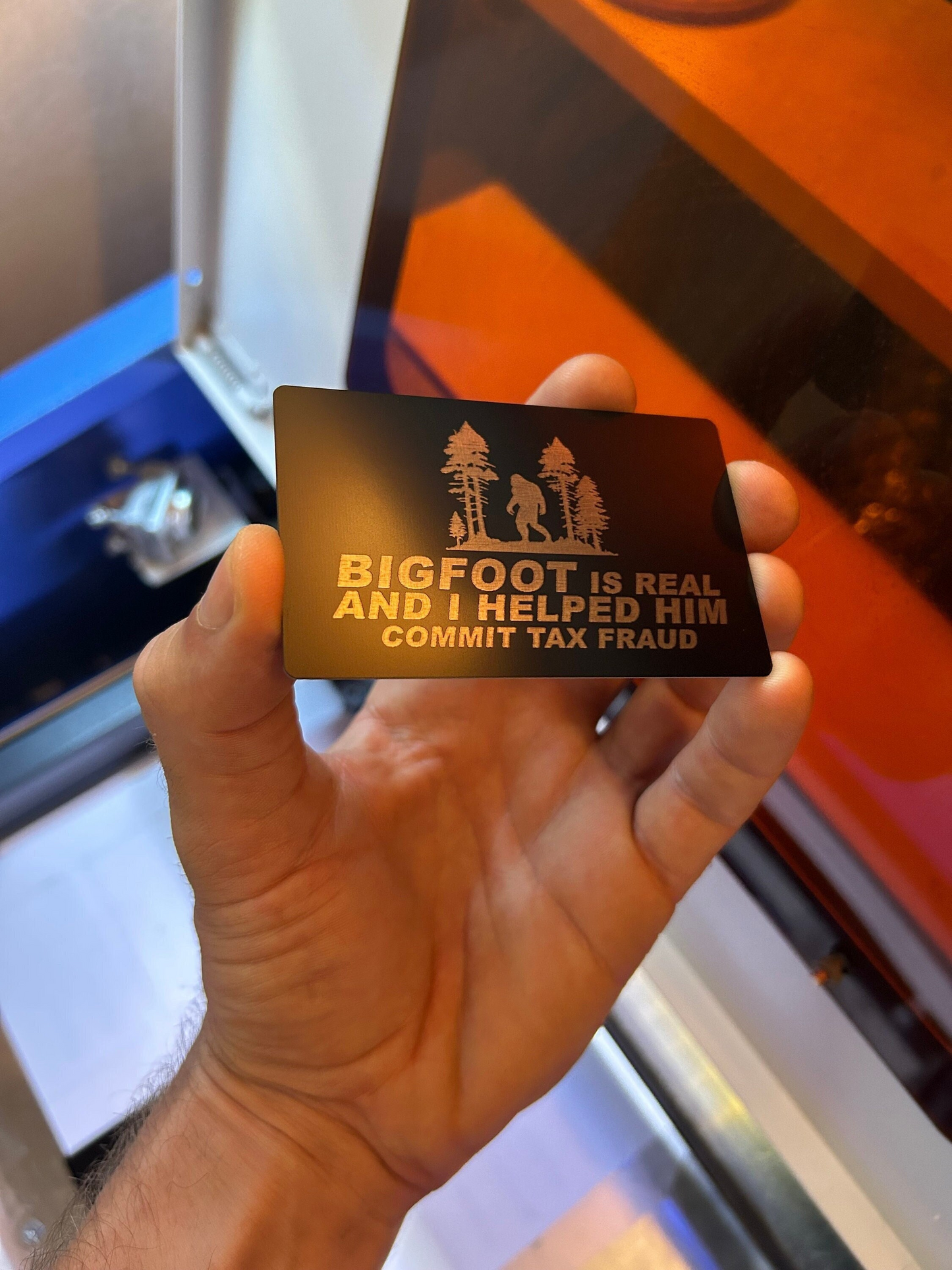 Hand holding a metal gift card with 'Bigfoot is real and I helped him commit tax fraud' text and a tree design.
