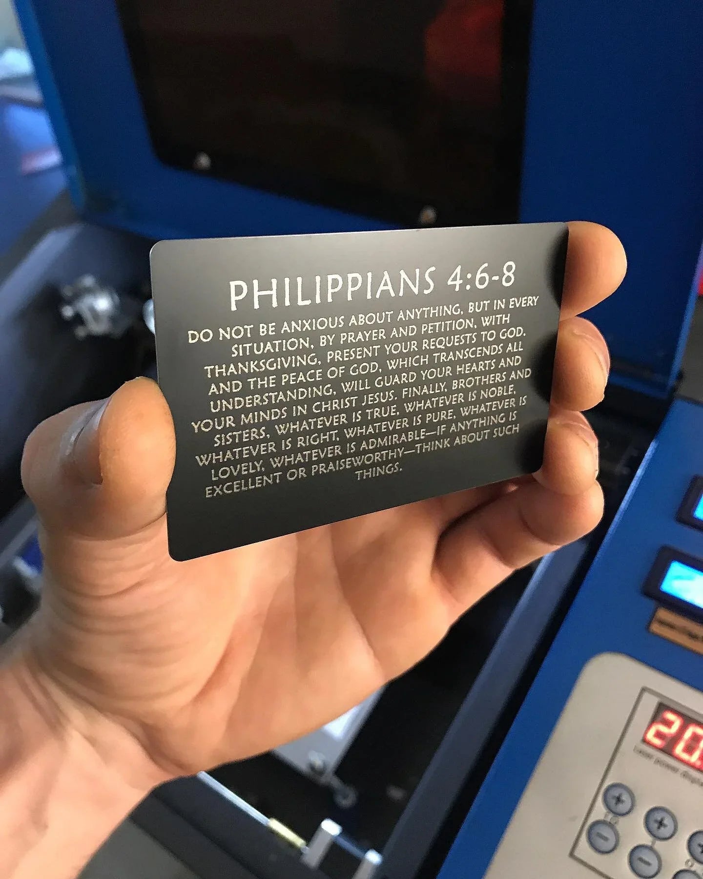 Hand holding a metal card with Philippians 4:6-8 text against a blue machine background