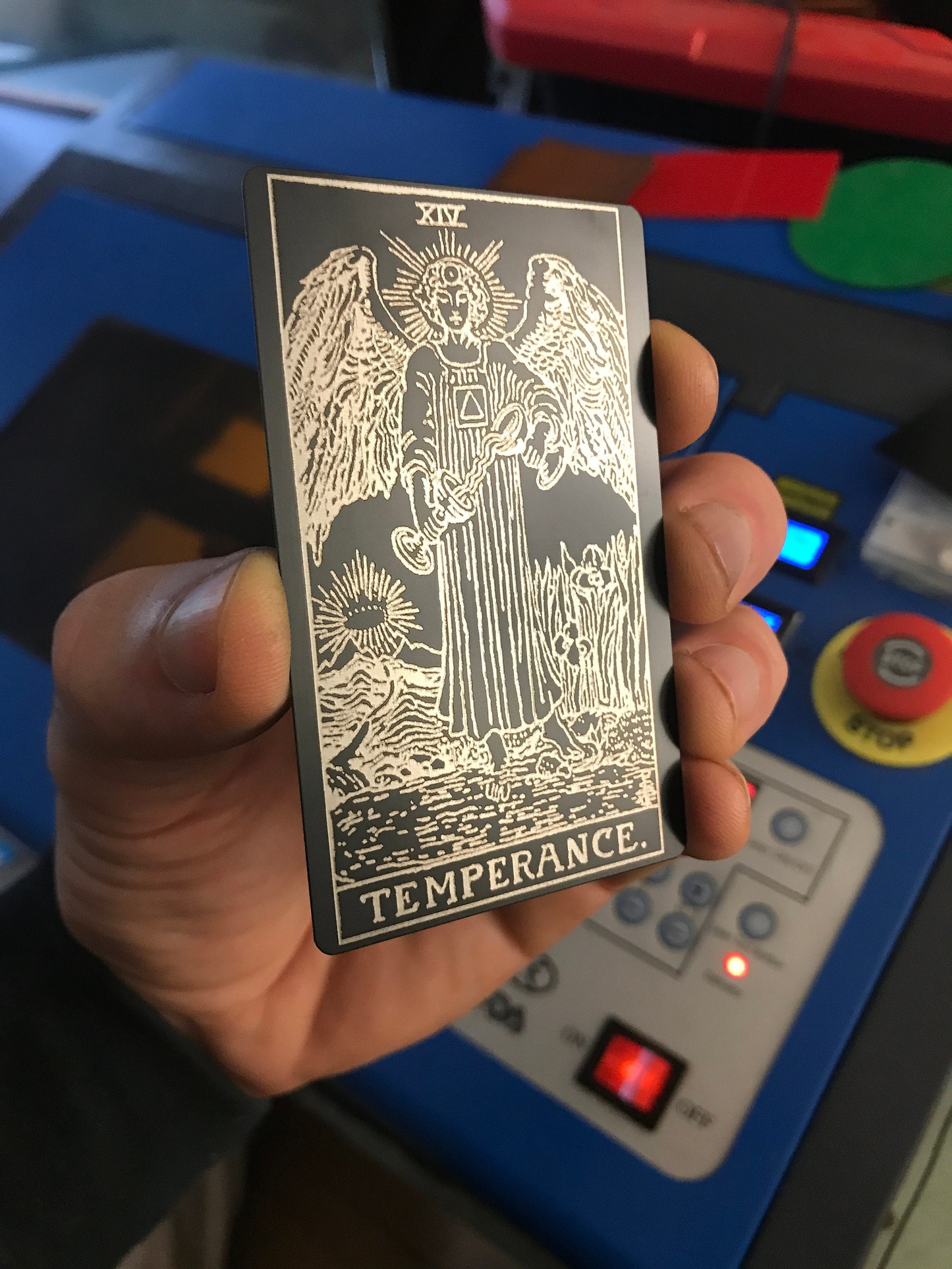 Hand holding a laser engraved metal tarot card titled 'Temperance' in front of a machine with buttons and lights.