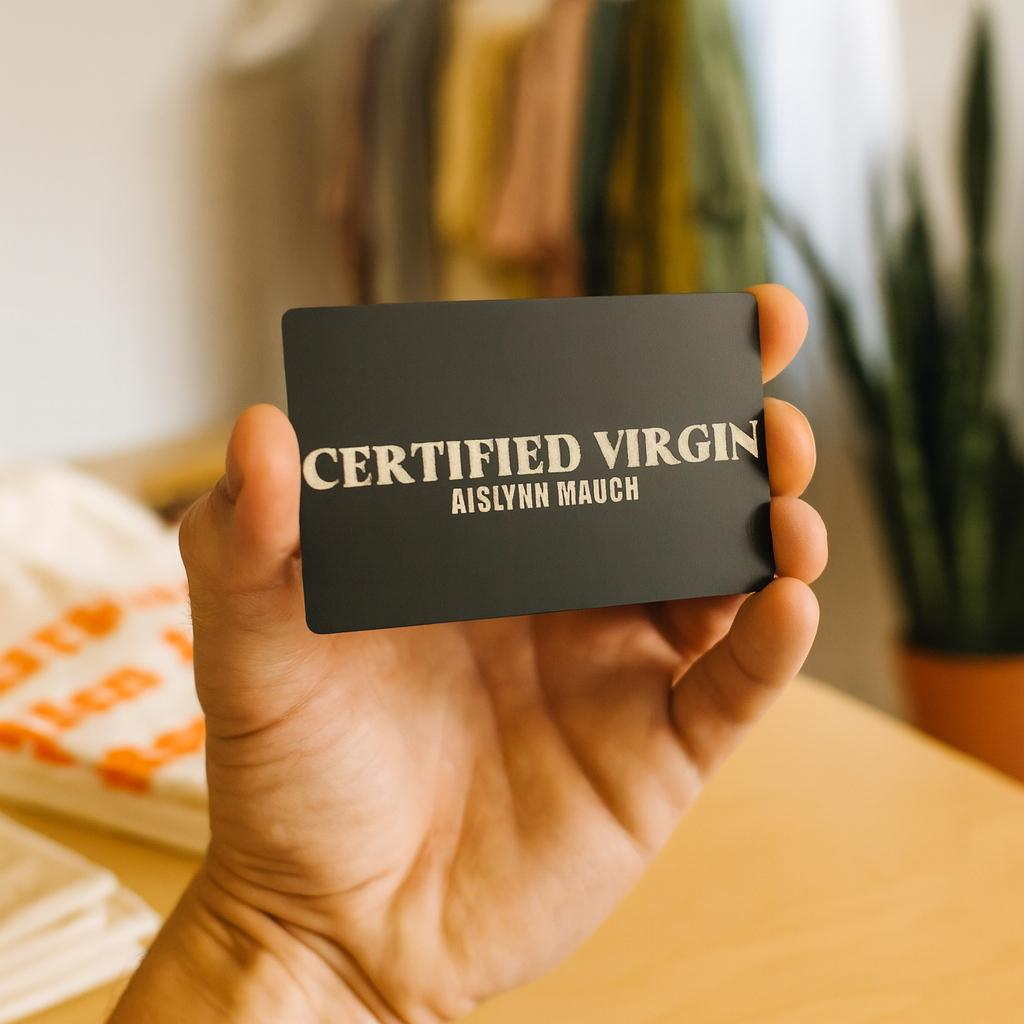 Hand holding a metal card with 'CERTIFIED VIRGIN AISLYNN MAUCH' customized text against a blurred indoor background.