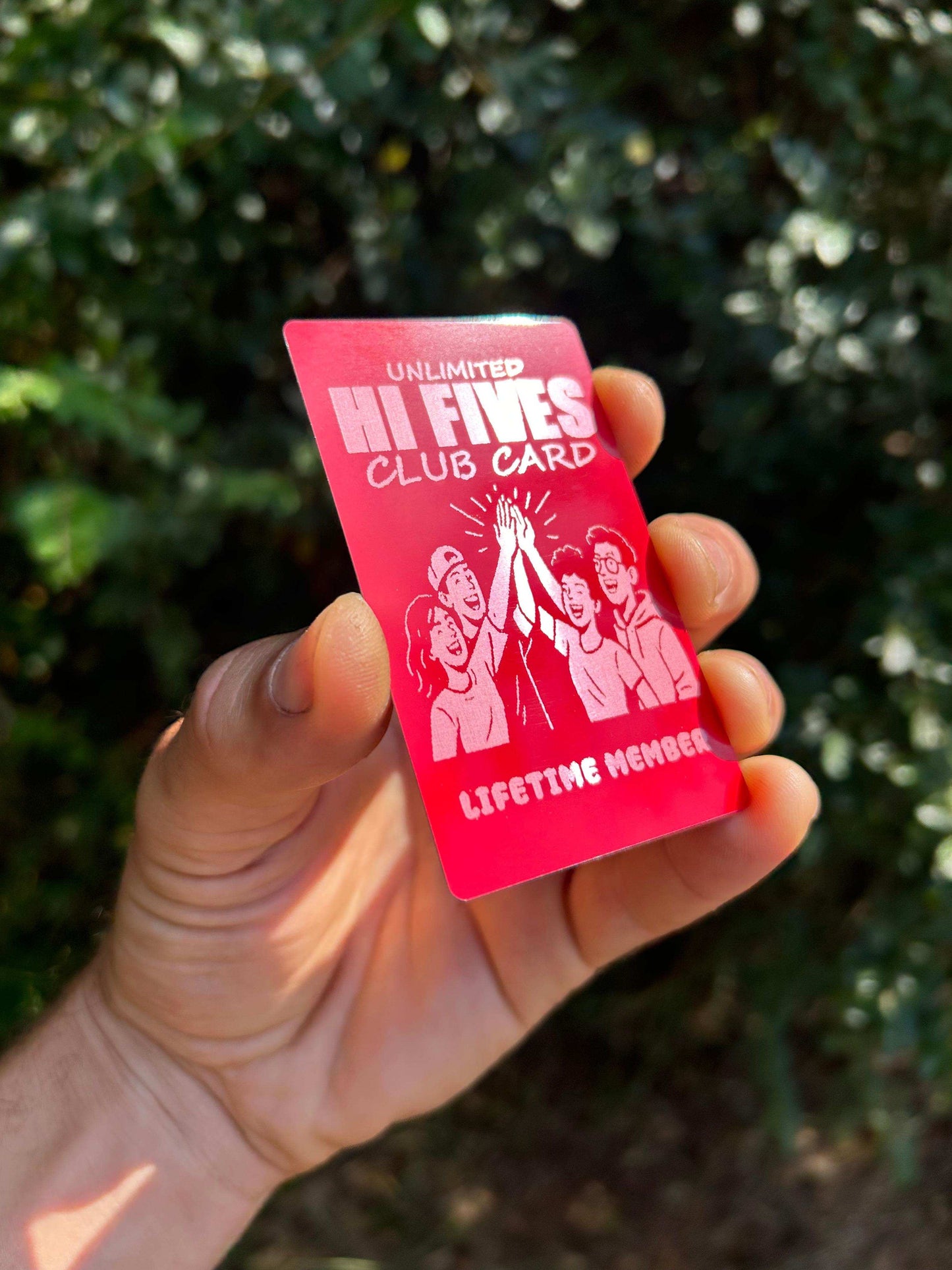 Hand holding a metal red/silver laser engraved 'Unlimited Hi Fives Club' card with an outdoor background