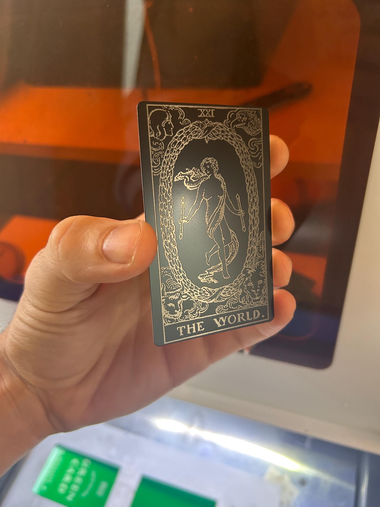 Hand holding a metal laser engraved black/silver tarot card titled 'The World' against a blurred background