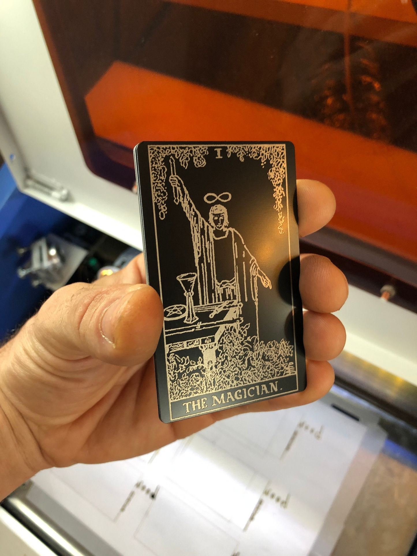 Metal laser engraved tarot Card with 'The Magician' design engraved, held by a hand in front of a blurred background
