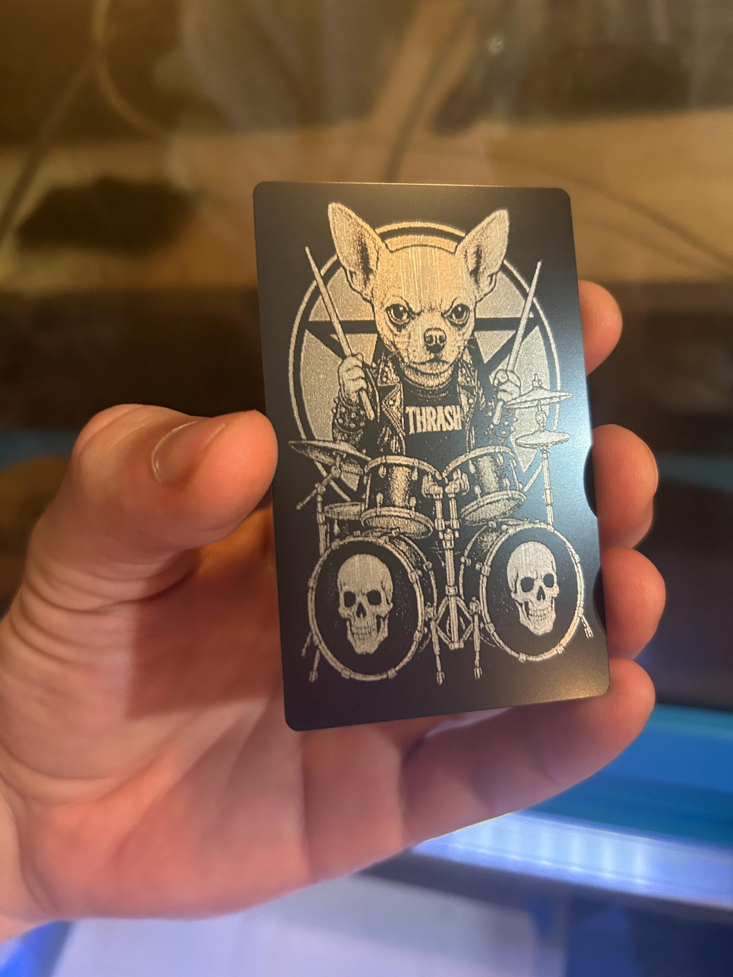 Hand holding a black laser engraved business card with a graphic of a dog playing drums and the word 'Thrash'.