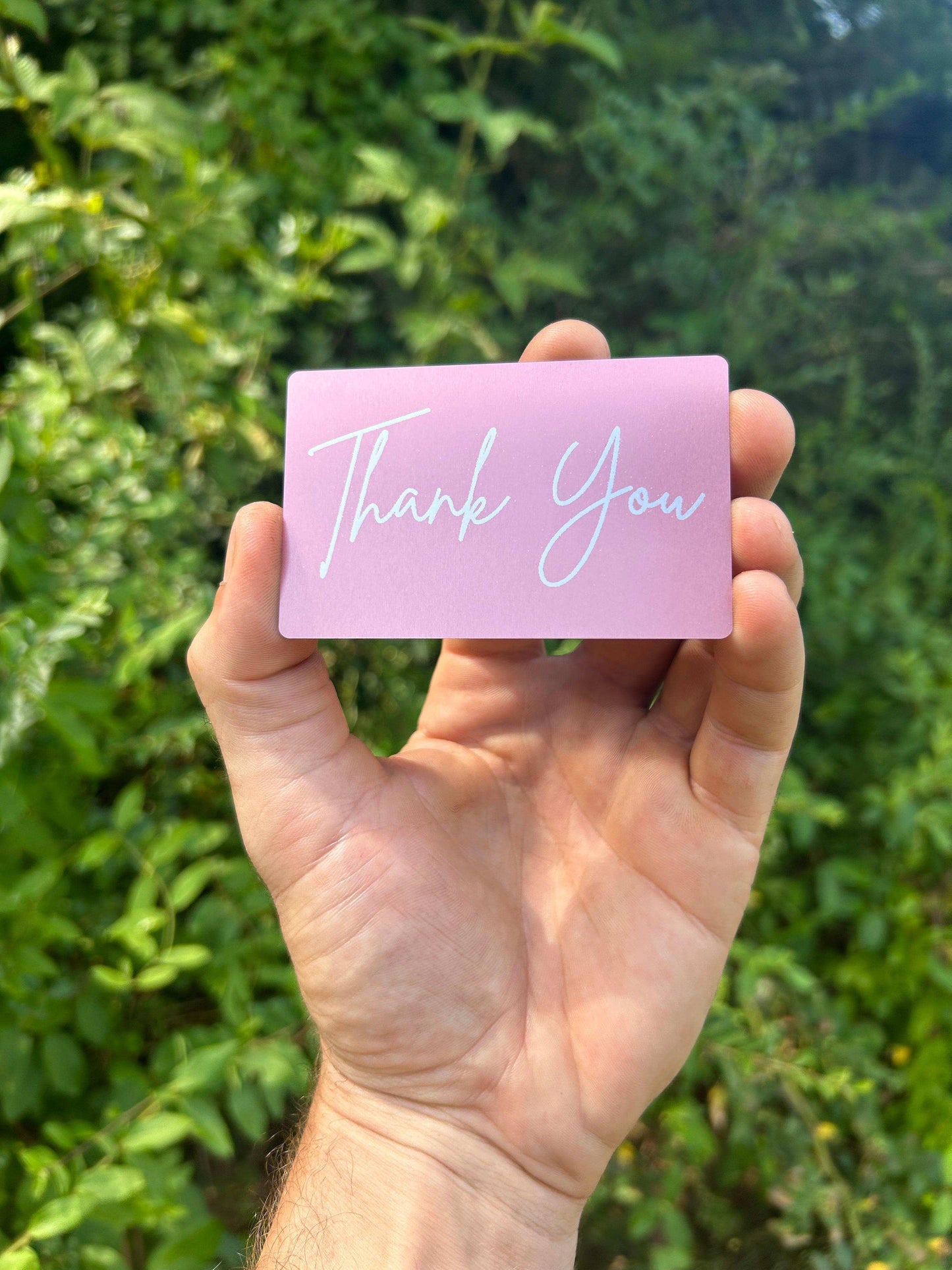 Hand holding a metal greeting card in rose gold with 'Thank You' text against a green leafy background