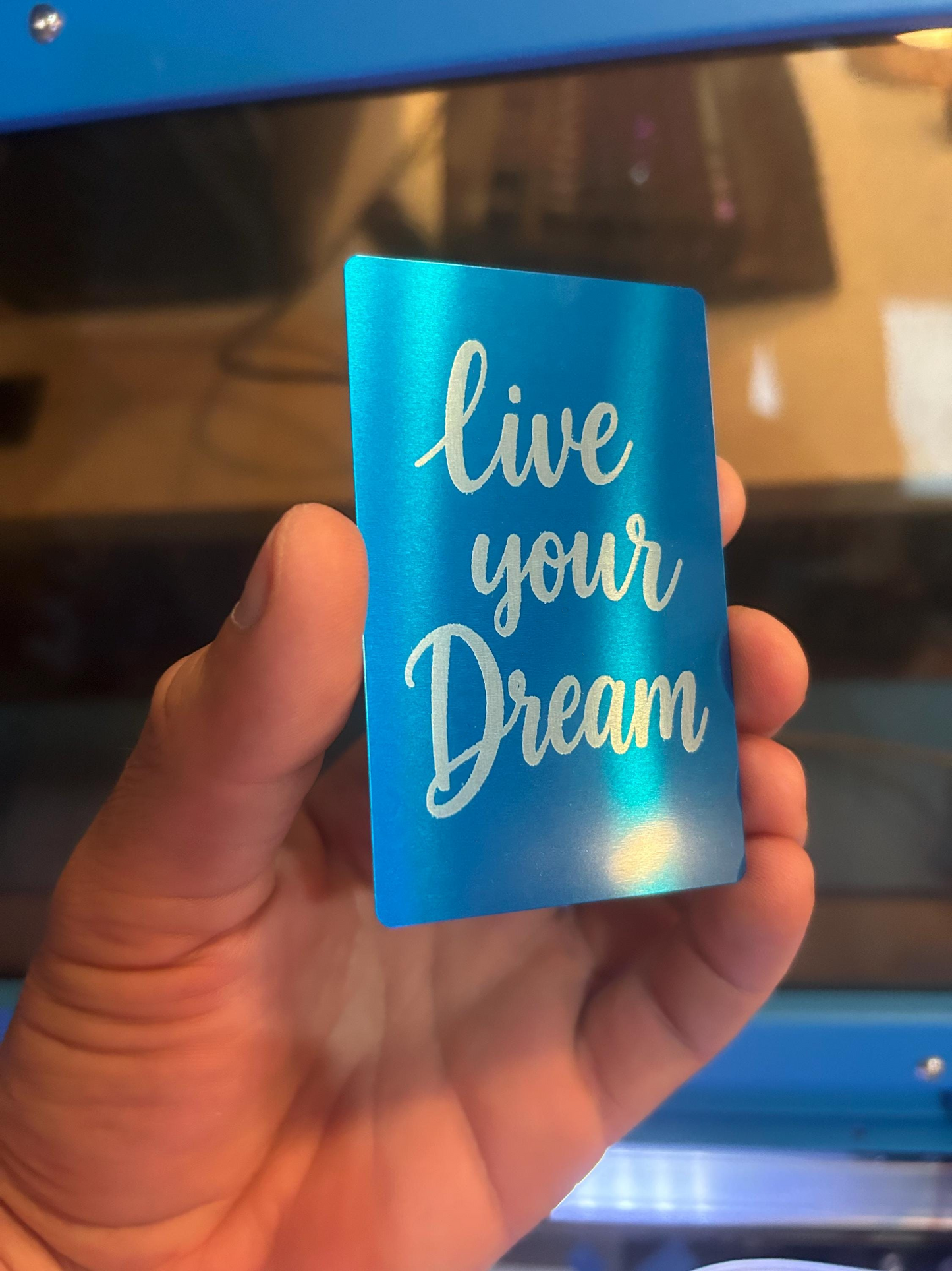 Hand presenting a blue and silver inspirational metal card – laser engraved “Live Your Dream” typography artwork
