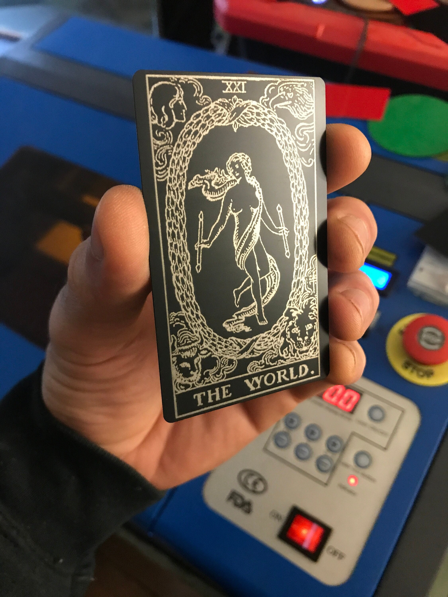 Hand holding a laser engraved metal tarot card titled 'The World' in front of a control panel.