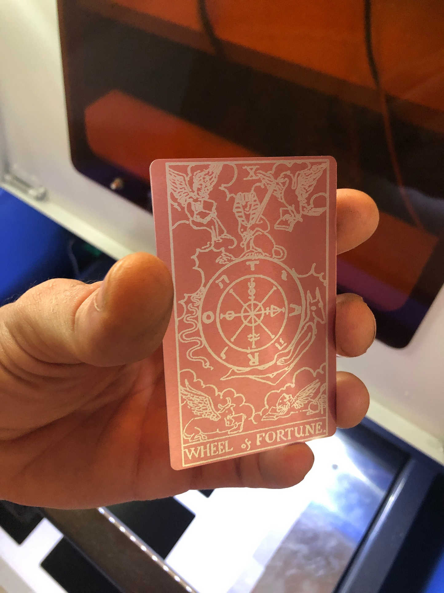 Hand holding a metal rose gold/silver tarot card with engraved Wheel of Fortune card designs and text, against a blurred indoor background.