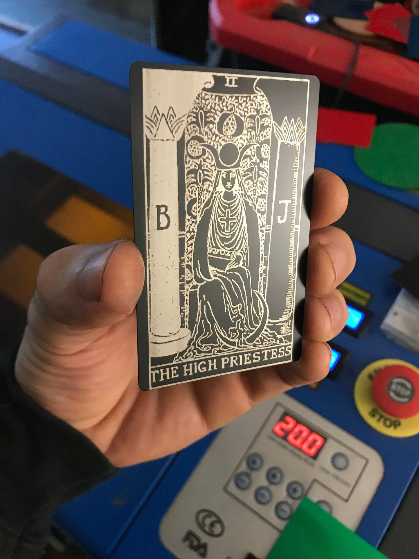 Hand holding a tarot card titled 'The High Priestess' laser engraved on metal against a colorful background.